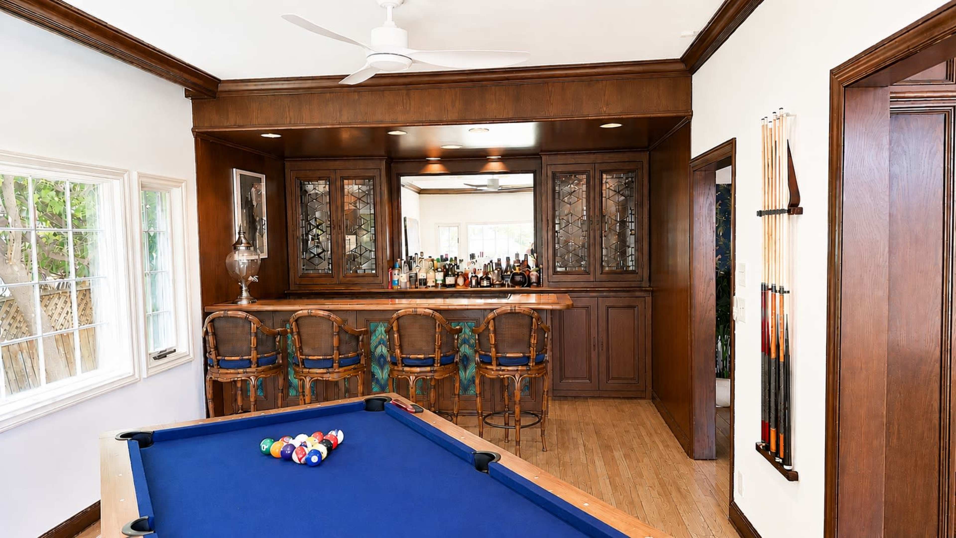 The image shows a room featuring a pool table in the foreground and a bar area with several chairs and shelves stocked with bottles in the background.