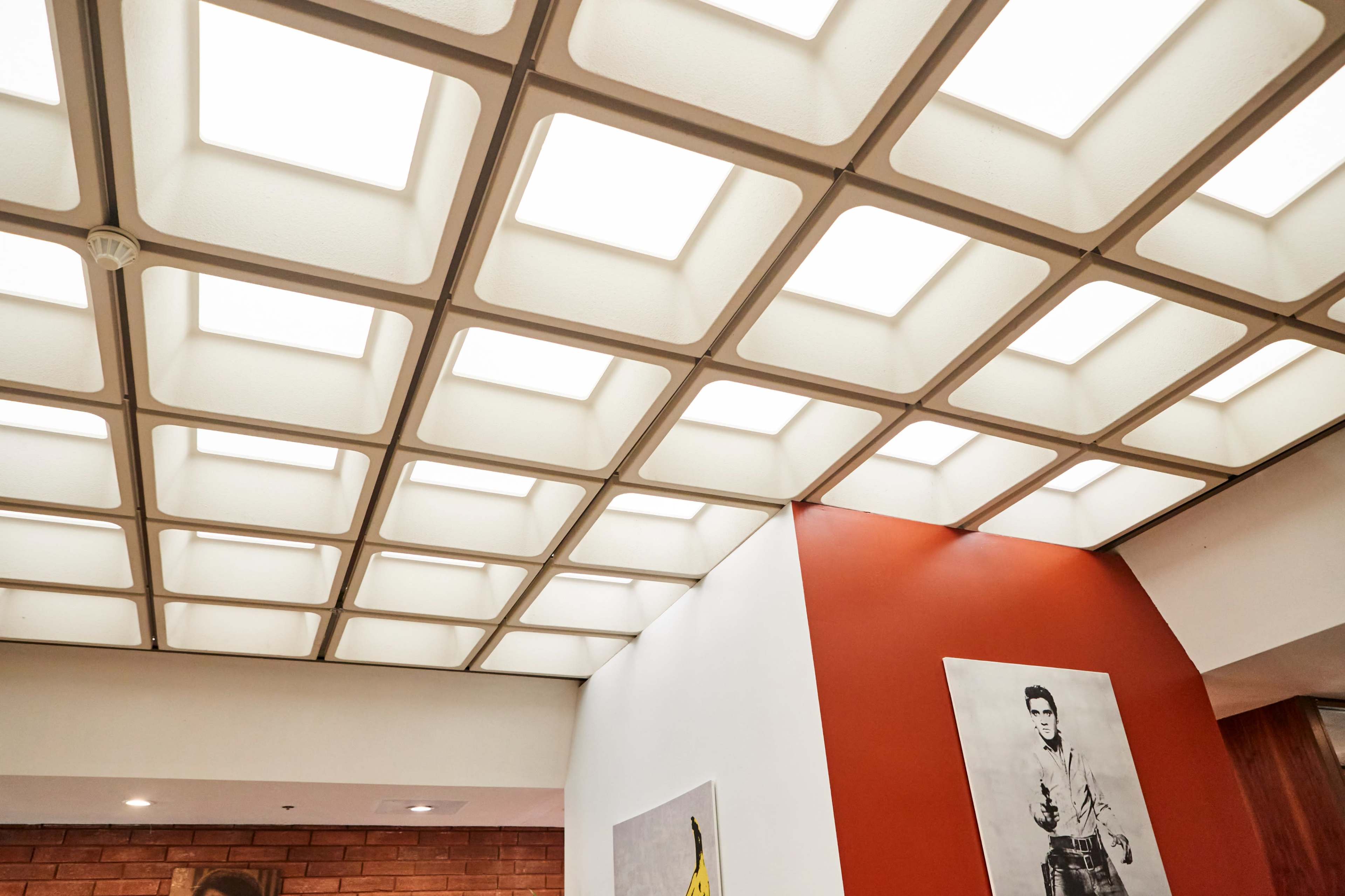 A ceiling with a grid pattern of translucent panels, complemented by a white wall and an orange accent wall featuring black-and-white artwork.