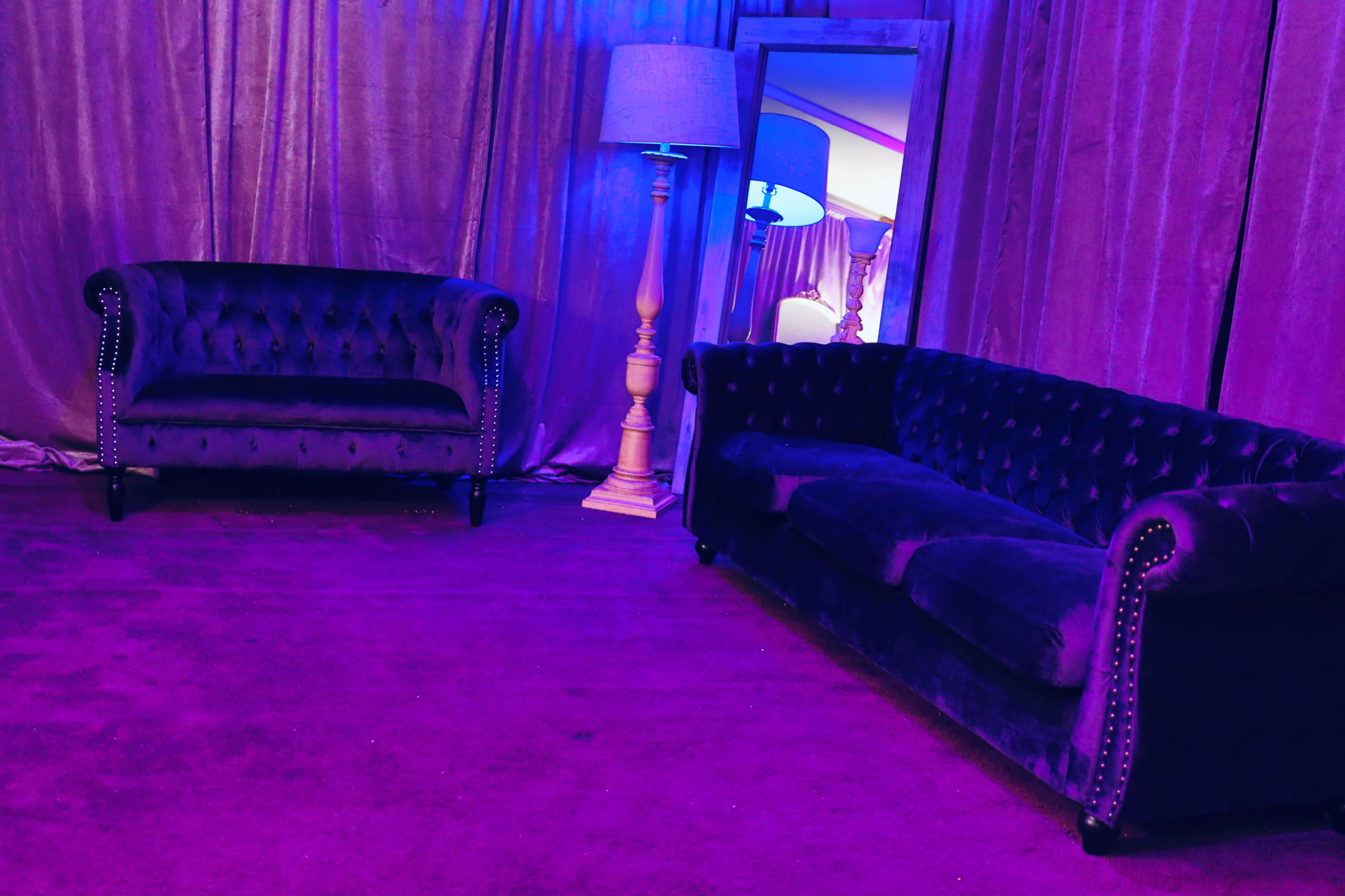 A purple-lit room features two velvet sofas, a lamp, and a mirror against a backdrop of draped fabric.