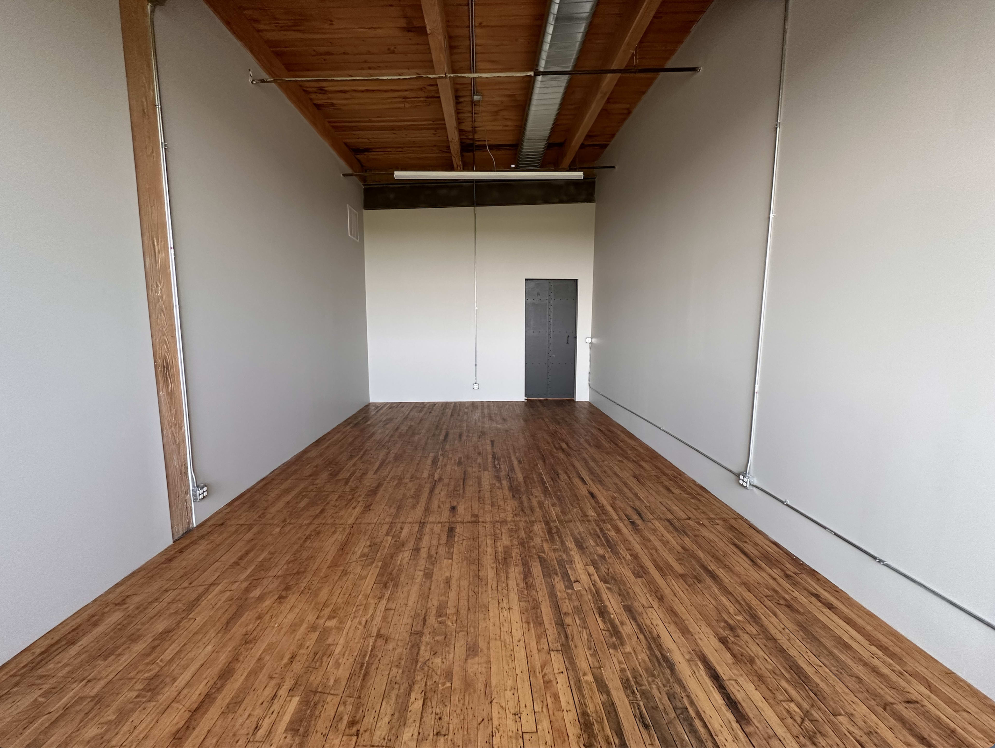 A long, empty room with wooden flooring, white walls, and a door at the far end.