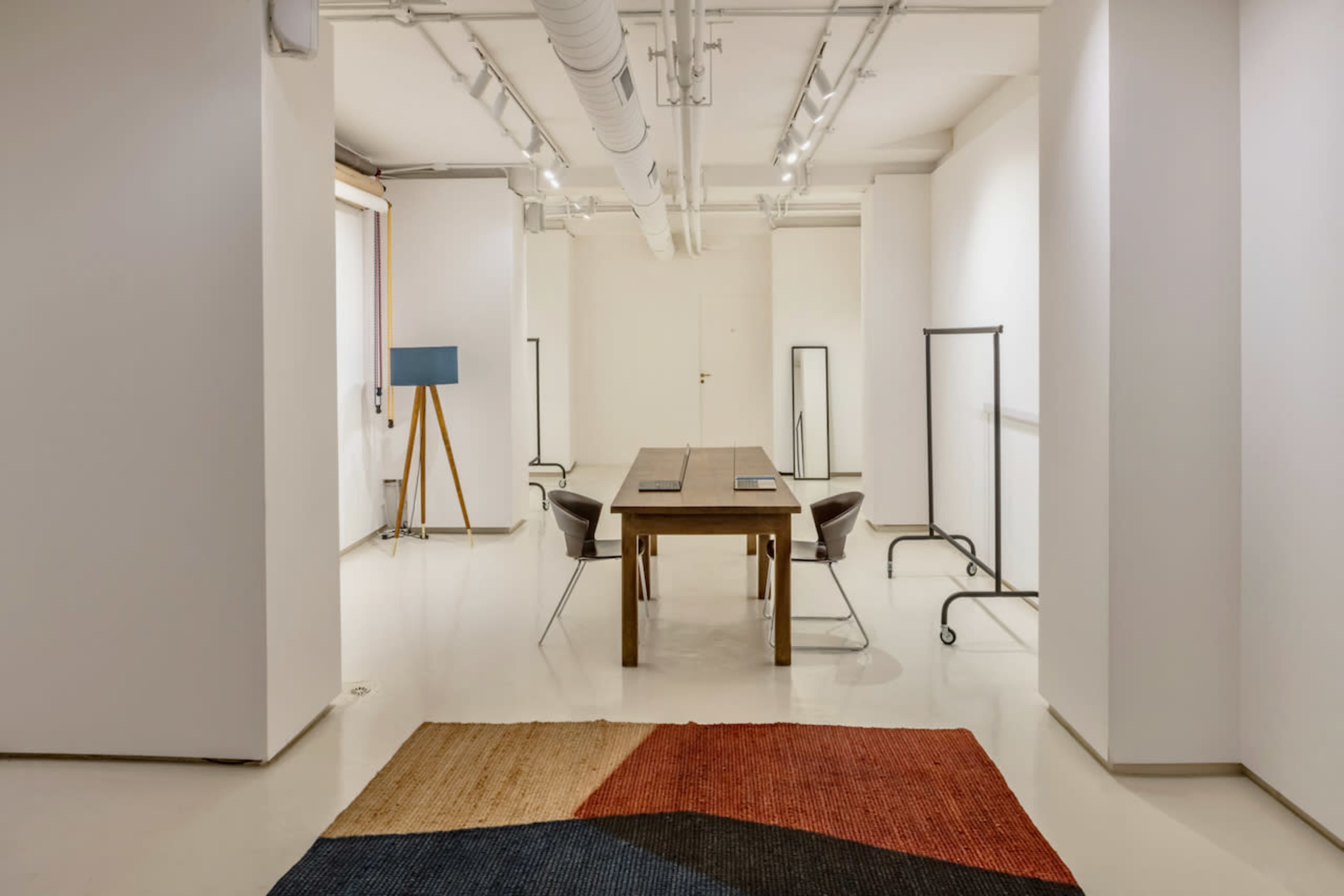 The image shows a minimalist office space featuring a long wooden table, two chairs, and a large rug, with white walls and ceiling lights.