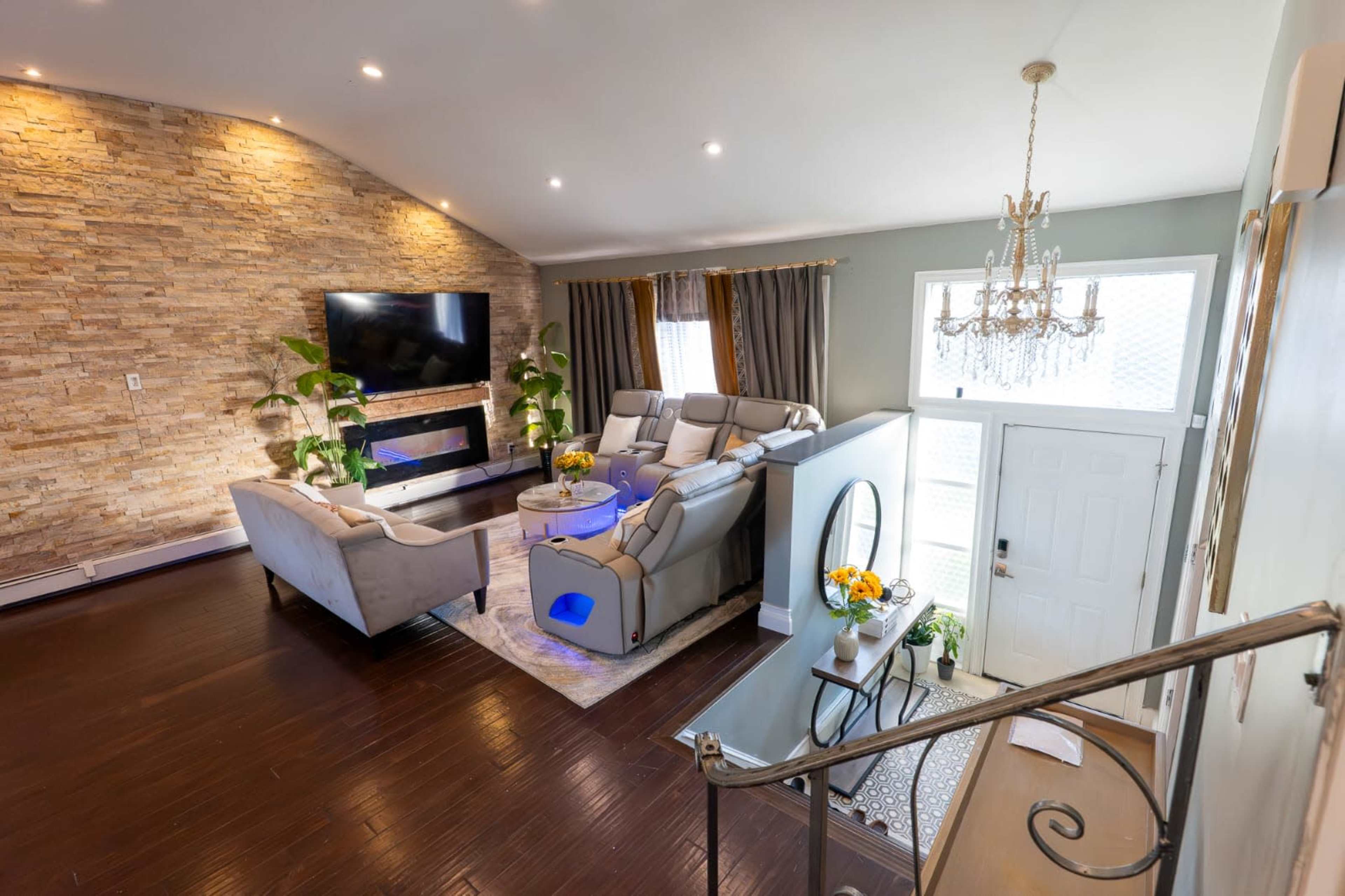The living room features a stone accent wall, modern seating arrangements, a large television mounted above a fireplace, and a chandelier overhead.