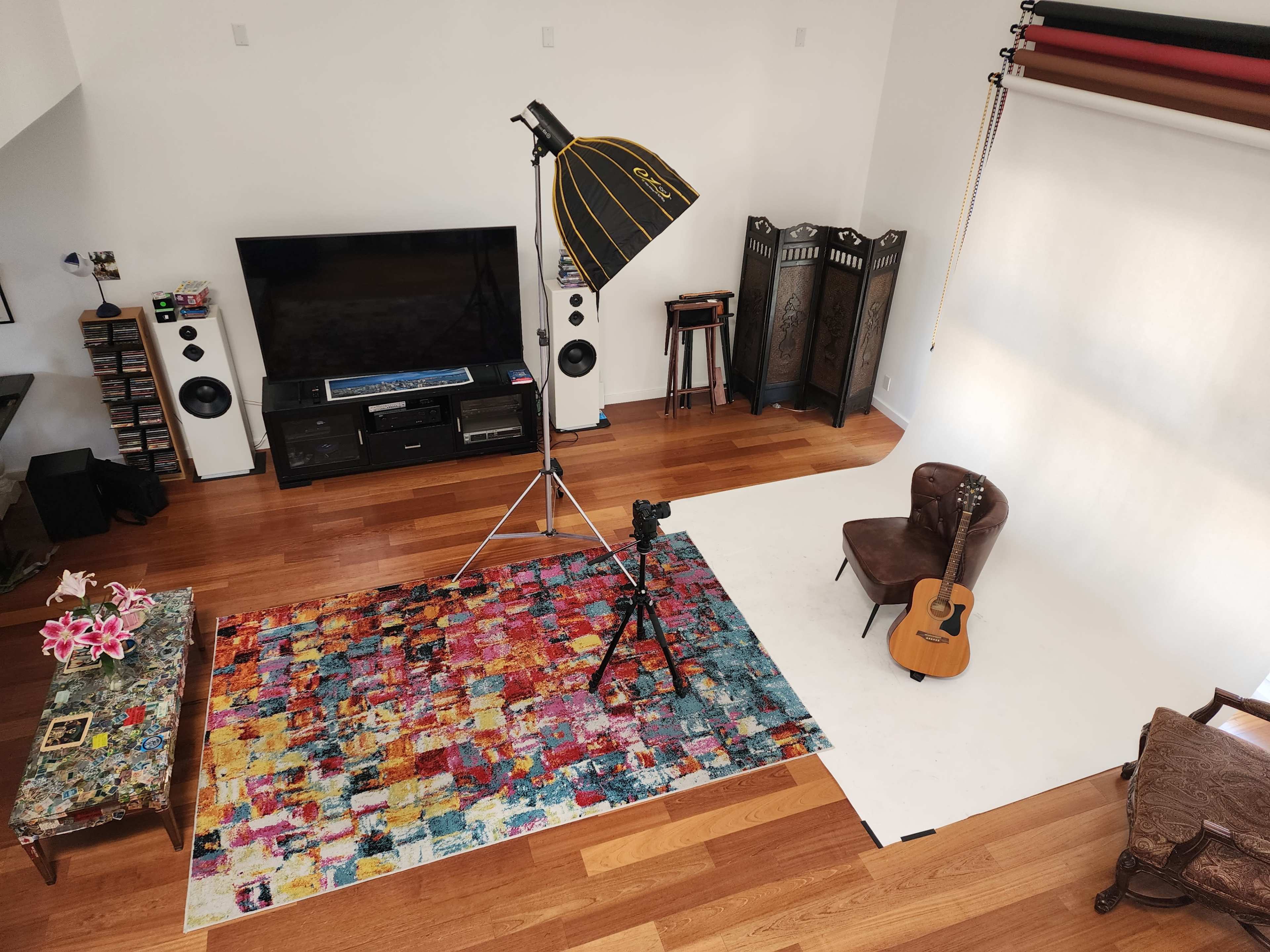 A well-lit photography studio with a tripod, guitar, and a colorful rug on wooden flooring.
