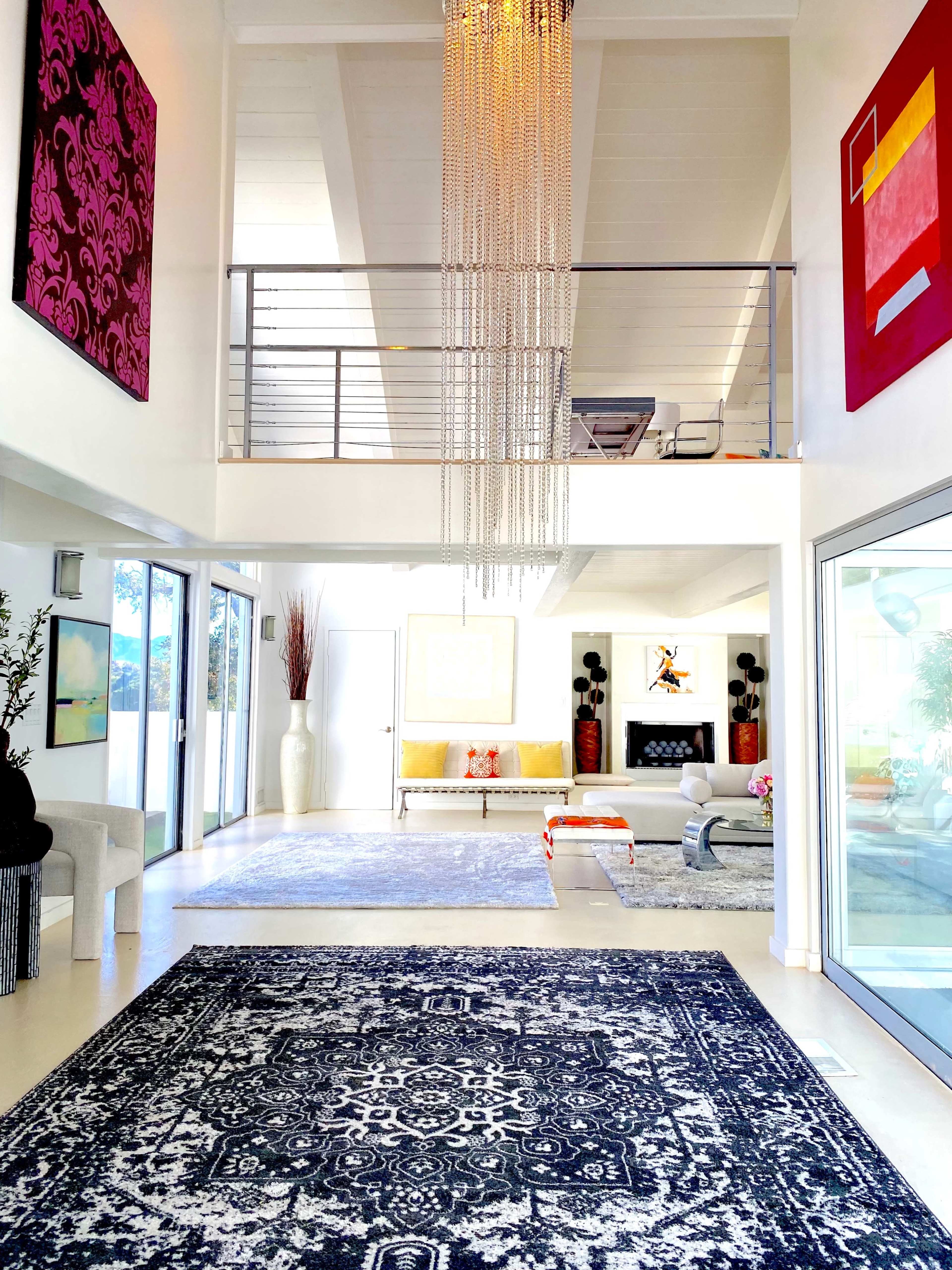 The image shows a spacious, modern interior with high ceilings, a chandelier, and colorful artwork on the walls, leading to a bright living area.