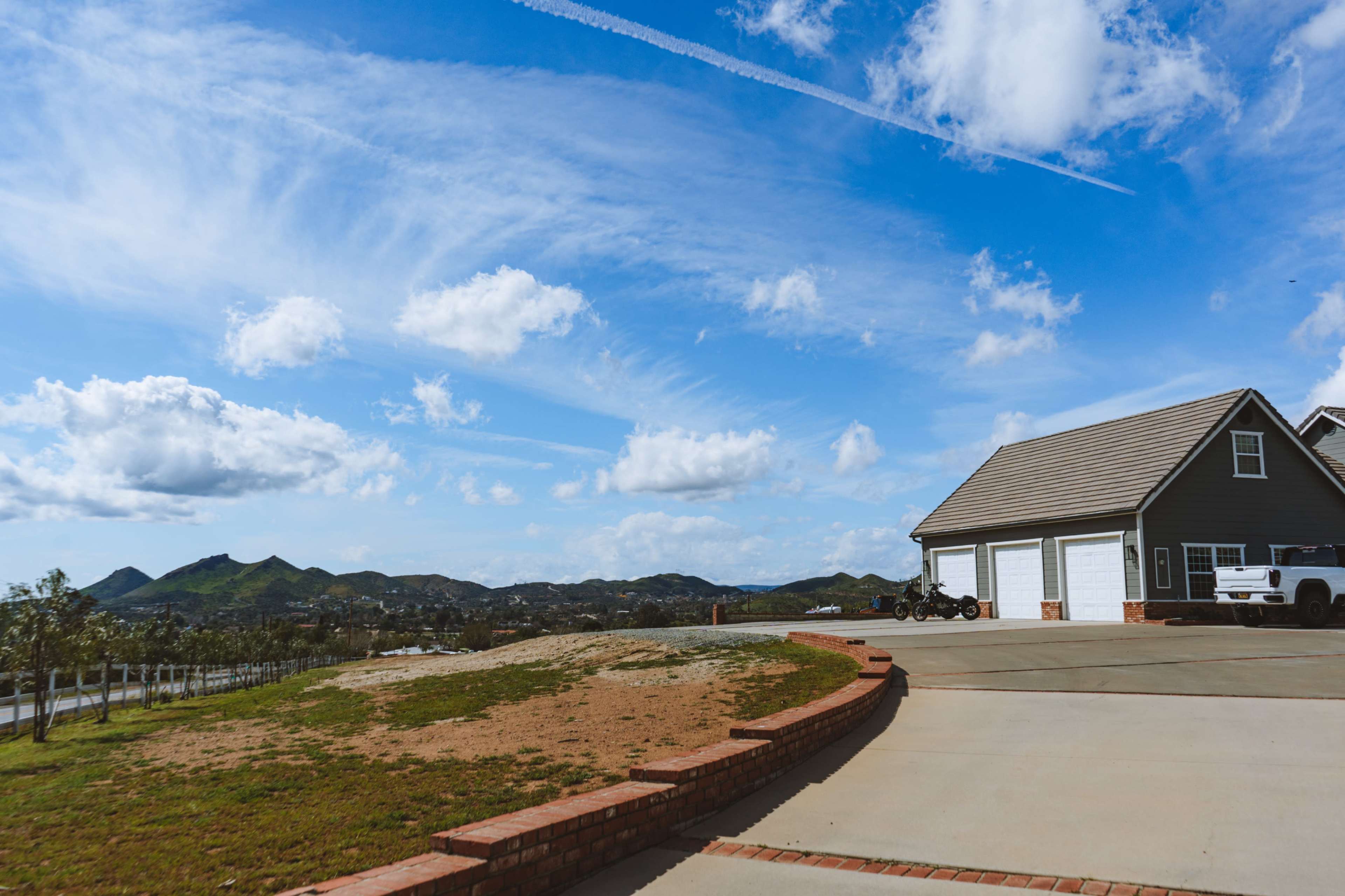 A house with a gray exterior and a three-car garage is set against a backdrop of rolling hills and a blue sky with scattered clouds.