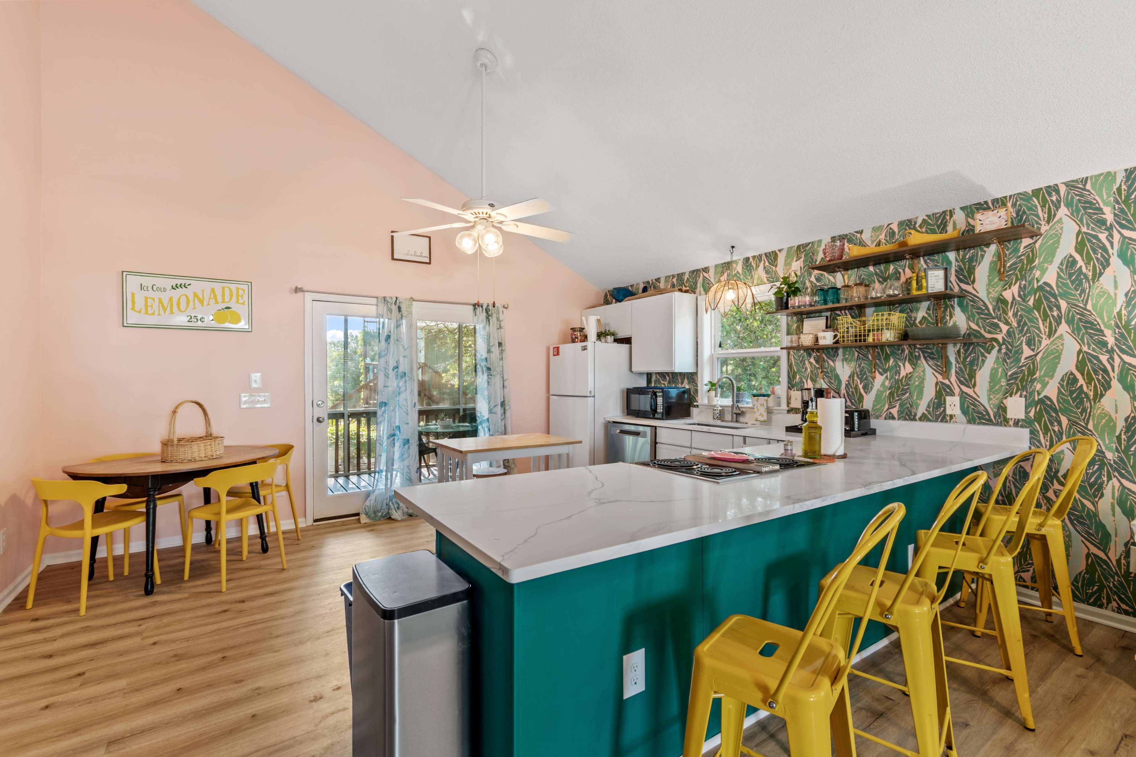 The image shows a vibrant kitchen with a modern island, yellow bar stools, and a dining area featuring a round table and colorful decor against a backdrop of pink and green walls.