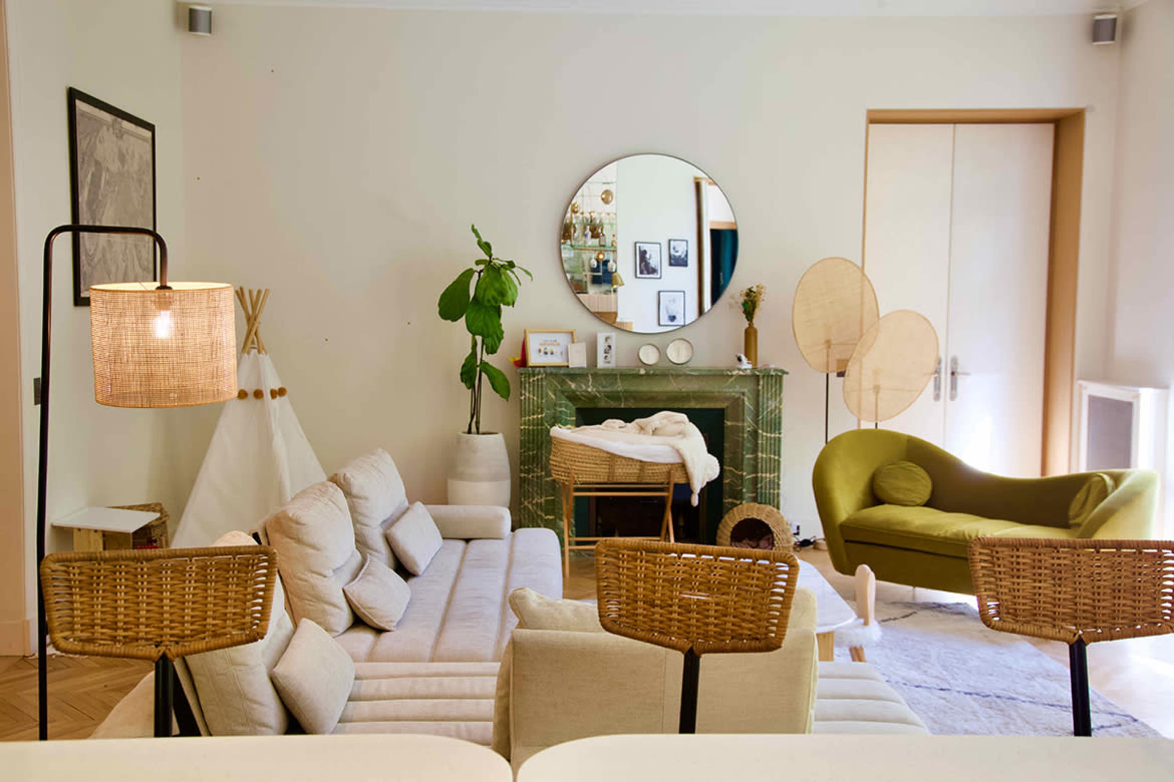 A cozy living room features a light-colored sofa, a green armchair, a decorative fireplace with a mirror above it, and a tall plant.