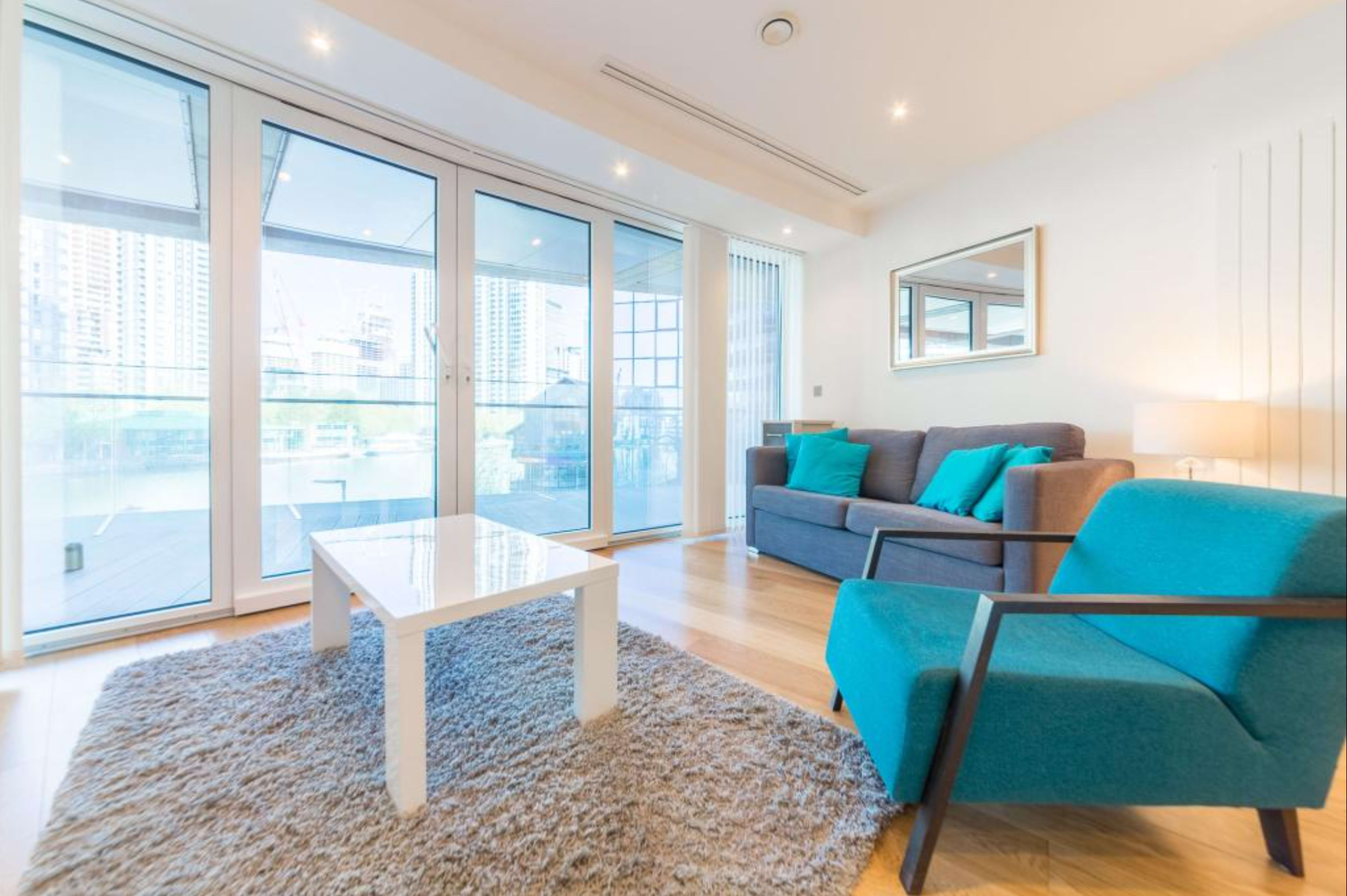 The image shows a bright living room with large glass doors, a gray sofa, a turquoise chair, and a white coffee table on a plush rug.