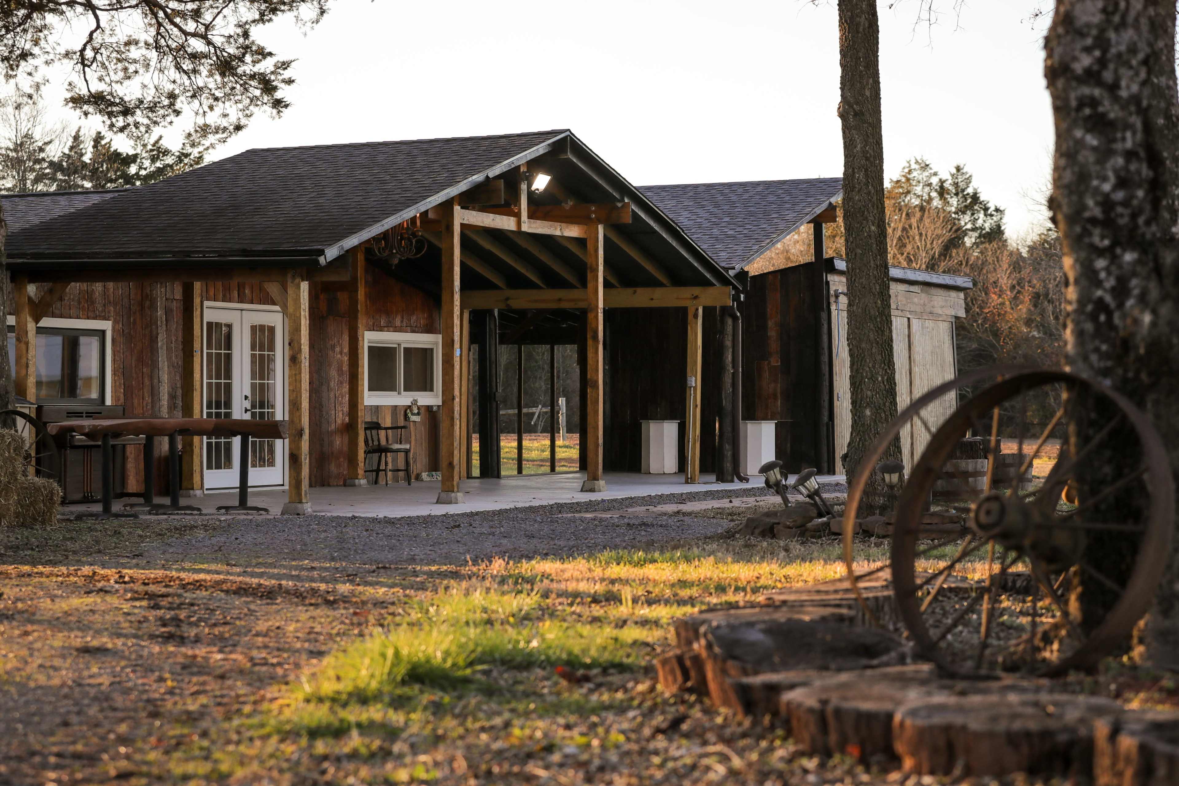 A rustic building with a wooden exterior and a porch is situated in a grassy area, surrounded by trees and logs.