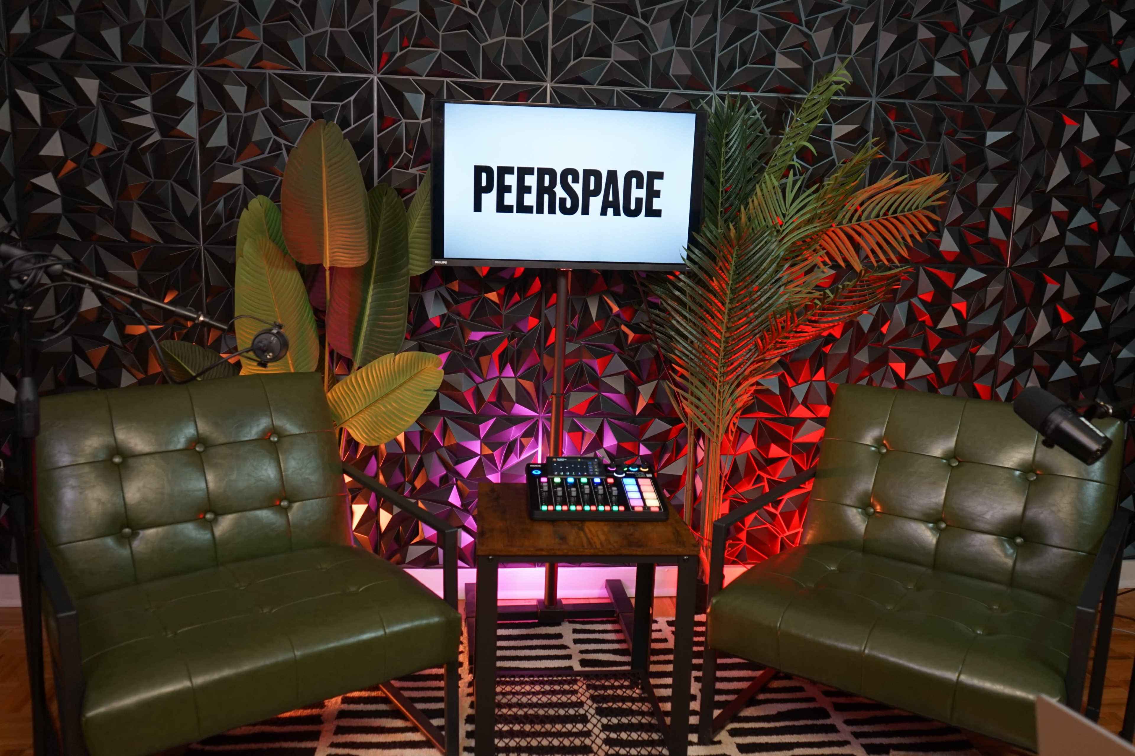 The image shows a small podcast or video production setup featuring two green chairs, a wooden table with a control panel, and a television screen displaying the word "PEERSPACE," all against a patterned black backdrop with colorful lighting.