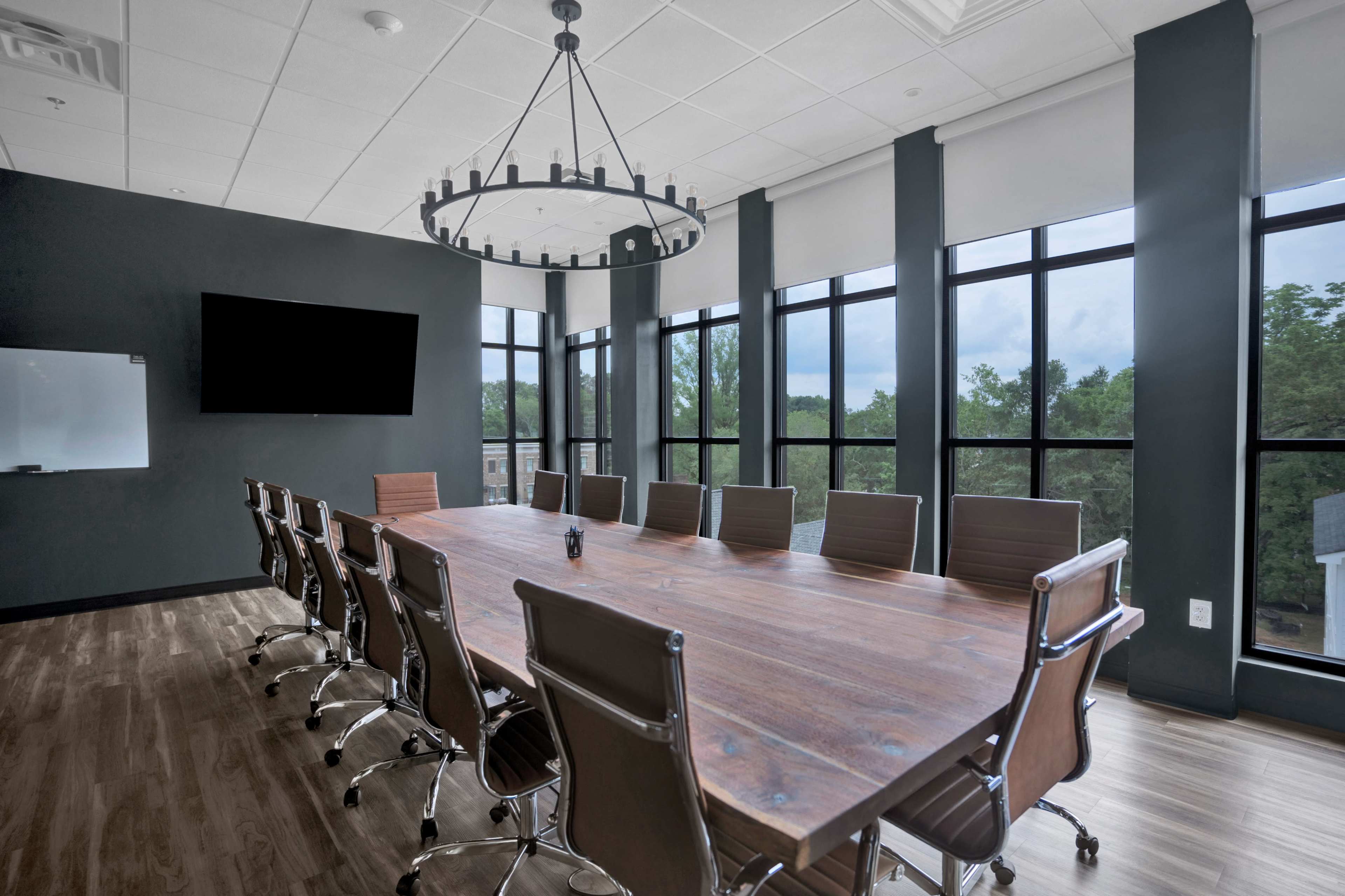 The image shows a conference room with a large wooden table surrounded by ergonomic chairs, large windows providing natural light, and a modern chandelier overhead.