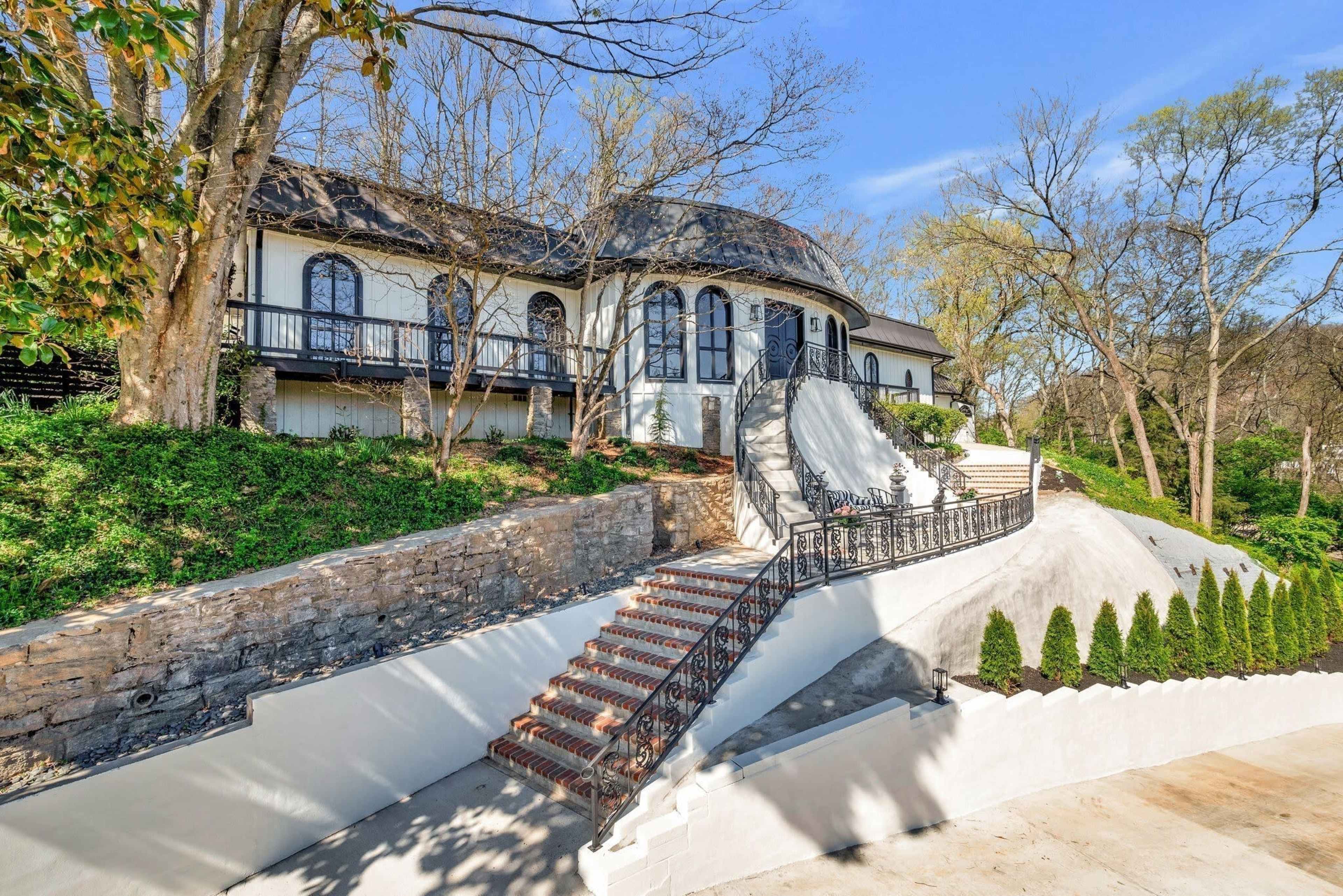 The image shows a large house with a curved roof, situated on a hillside with a landscaped pathway leading up to a set of stairs.