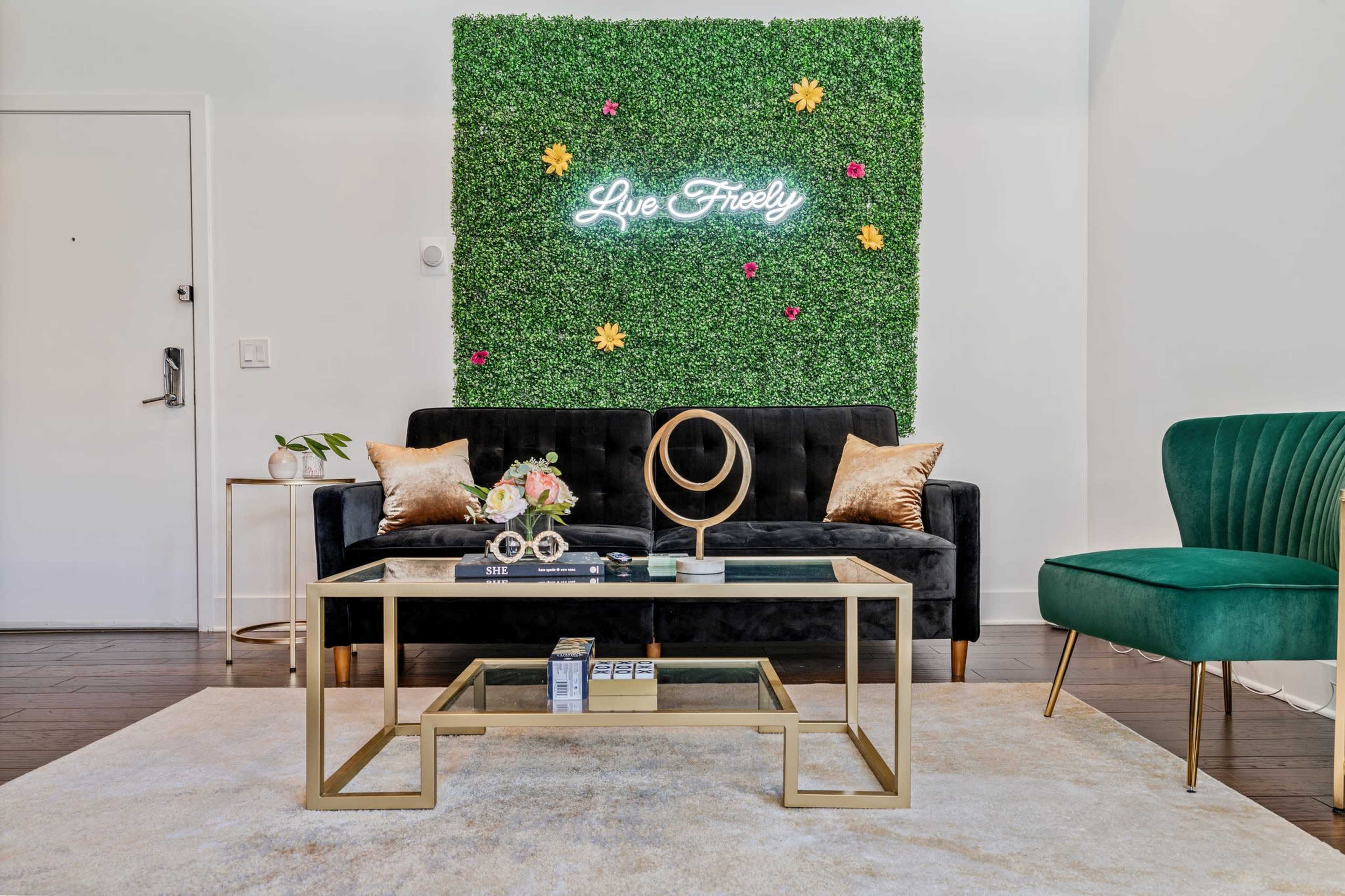 A cozy living room features a green wall with a neon sign reading "Live Freely," a black sofa, a glass coffee table, and a green accent chair.