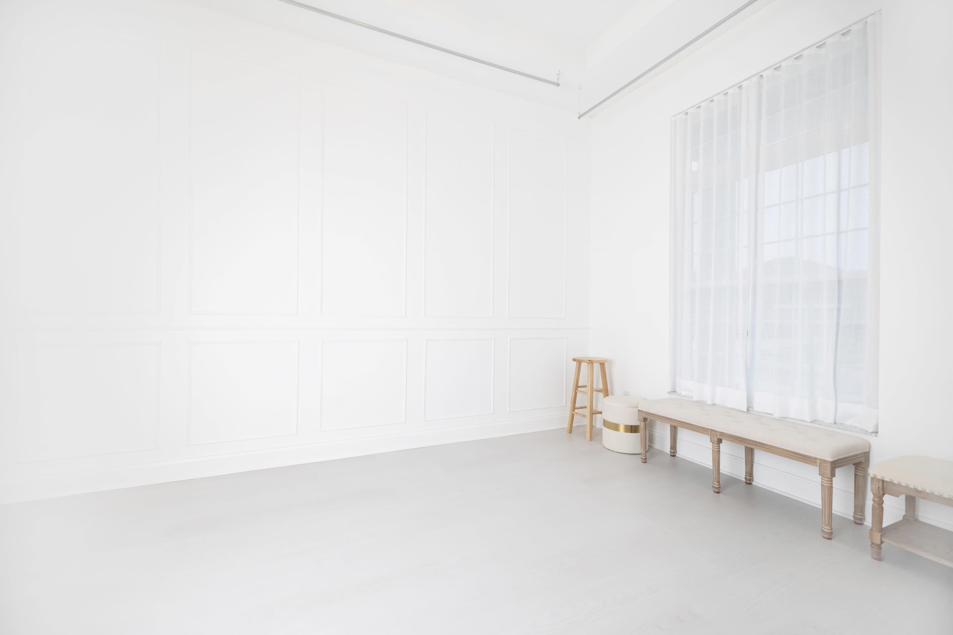The image shows a minimalist room with white walls, a large window, a wooden bench, a small stool, and a light-colored floor.