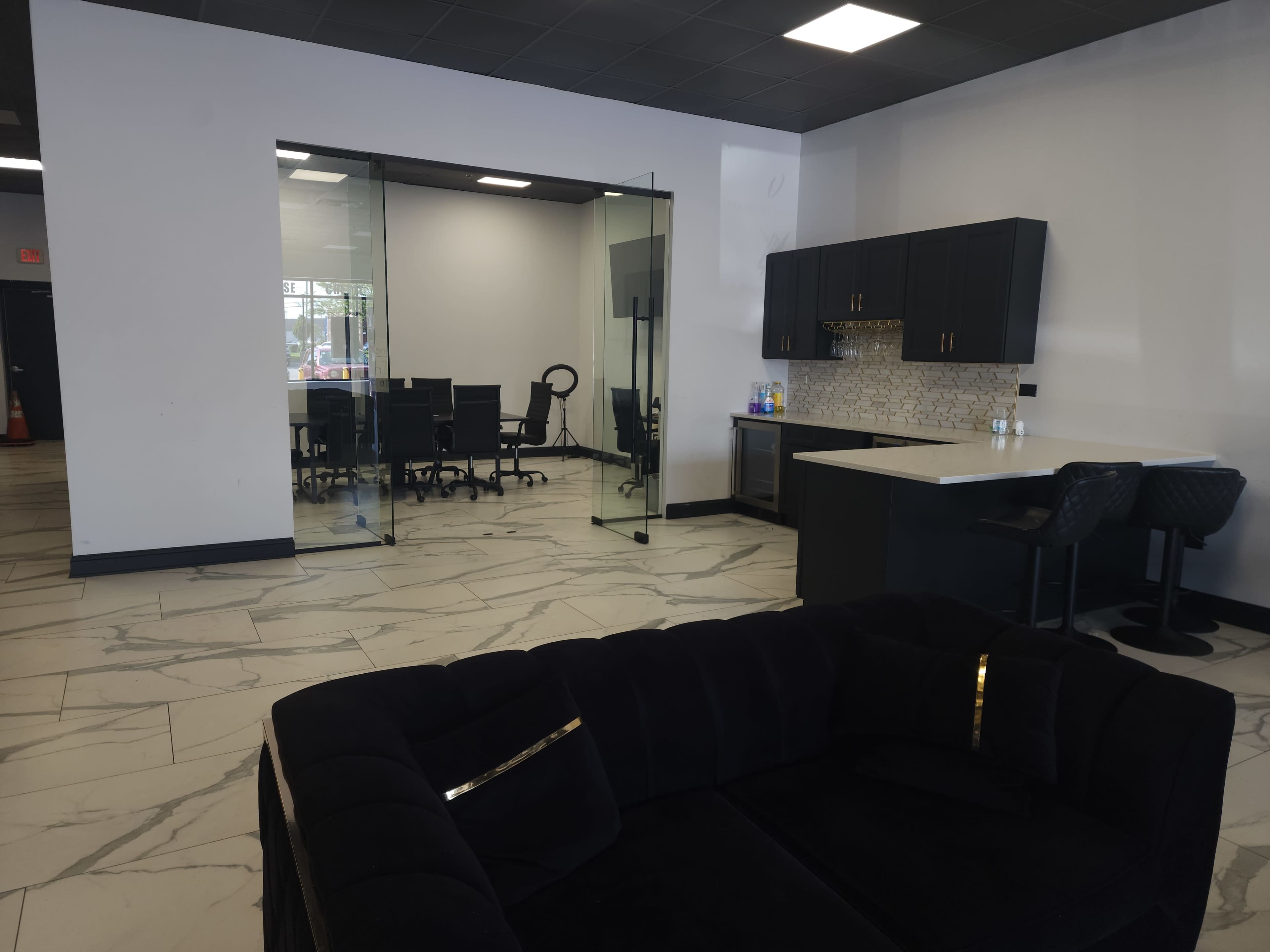 The image shows a modern office space with a black couch in the foreground, a glass-walled meeting room in the background, and a kitchenette with black cabinetry.