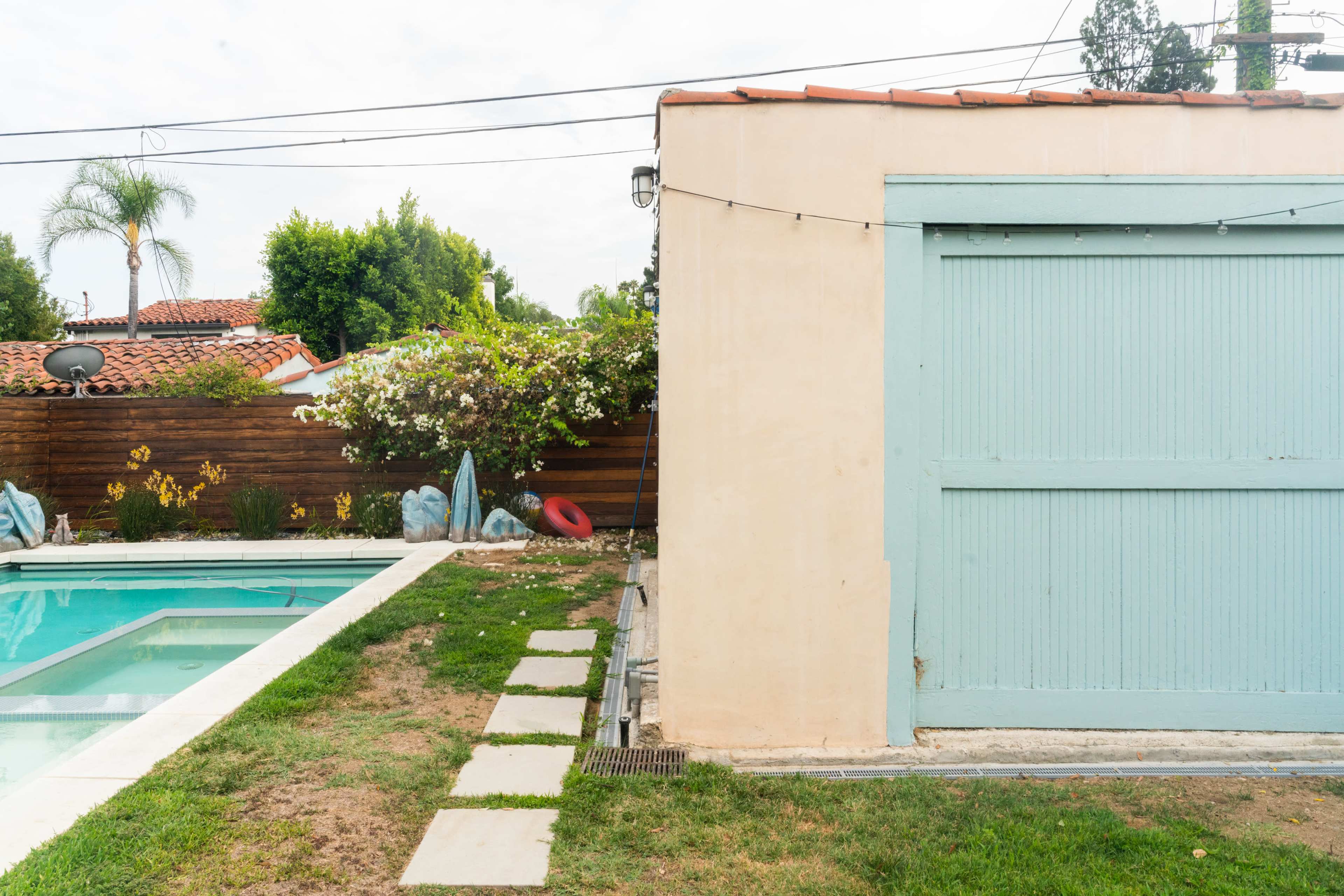 A light blue shed is situated next to a pool, with a grassy area and a wooden fence in the background.