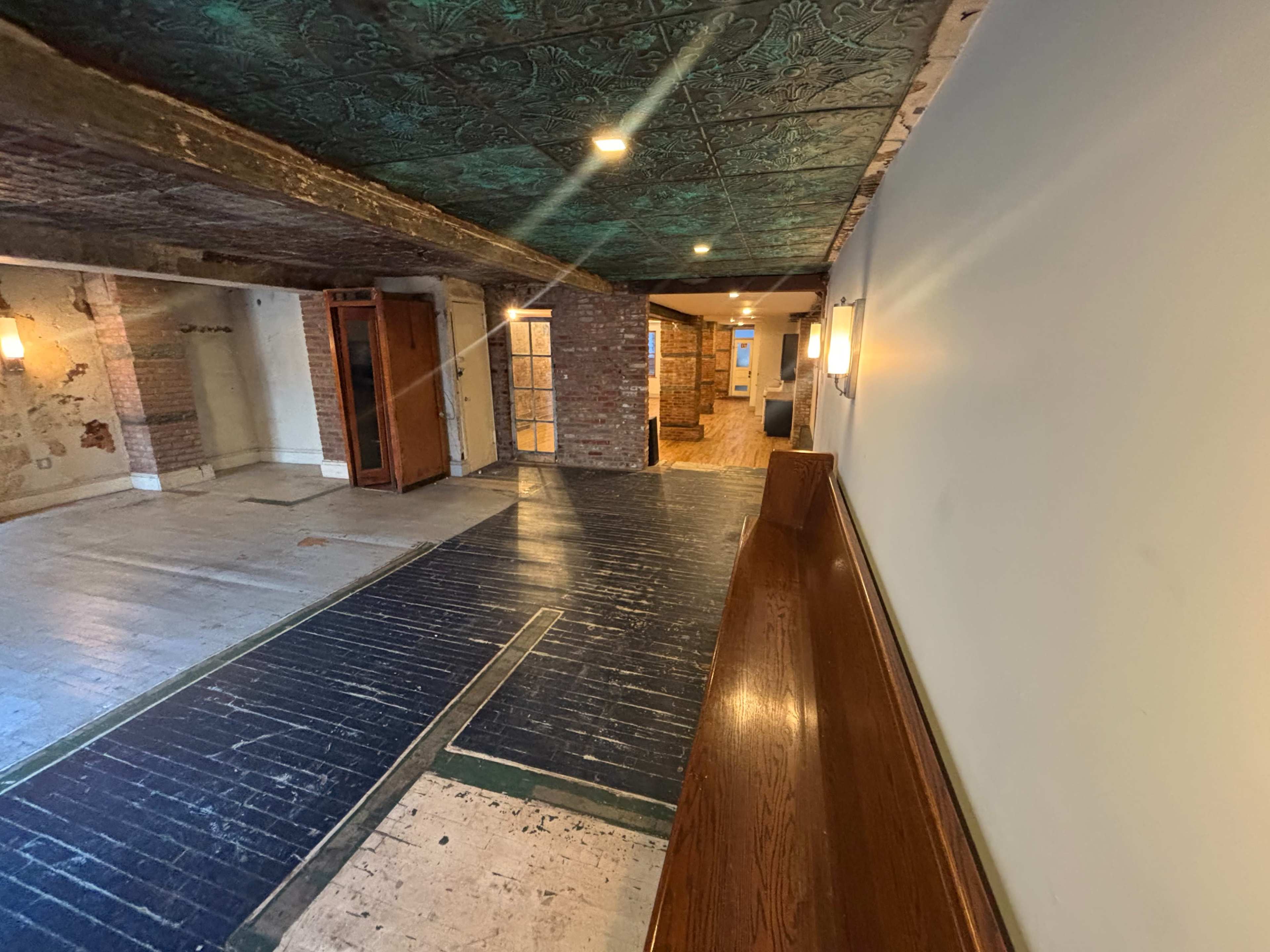 The image shows an interior space with a wooden bench on the left, exposed brick walls, and a mix of old and new flooring leading to a doorway at the far end.