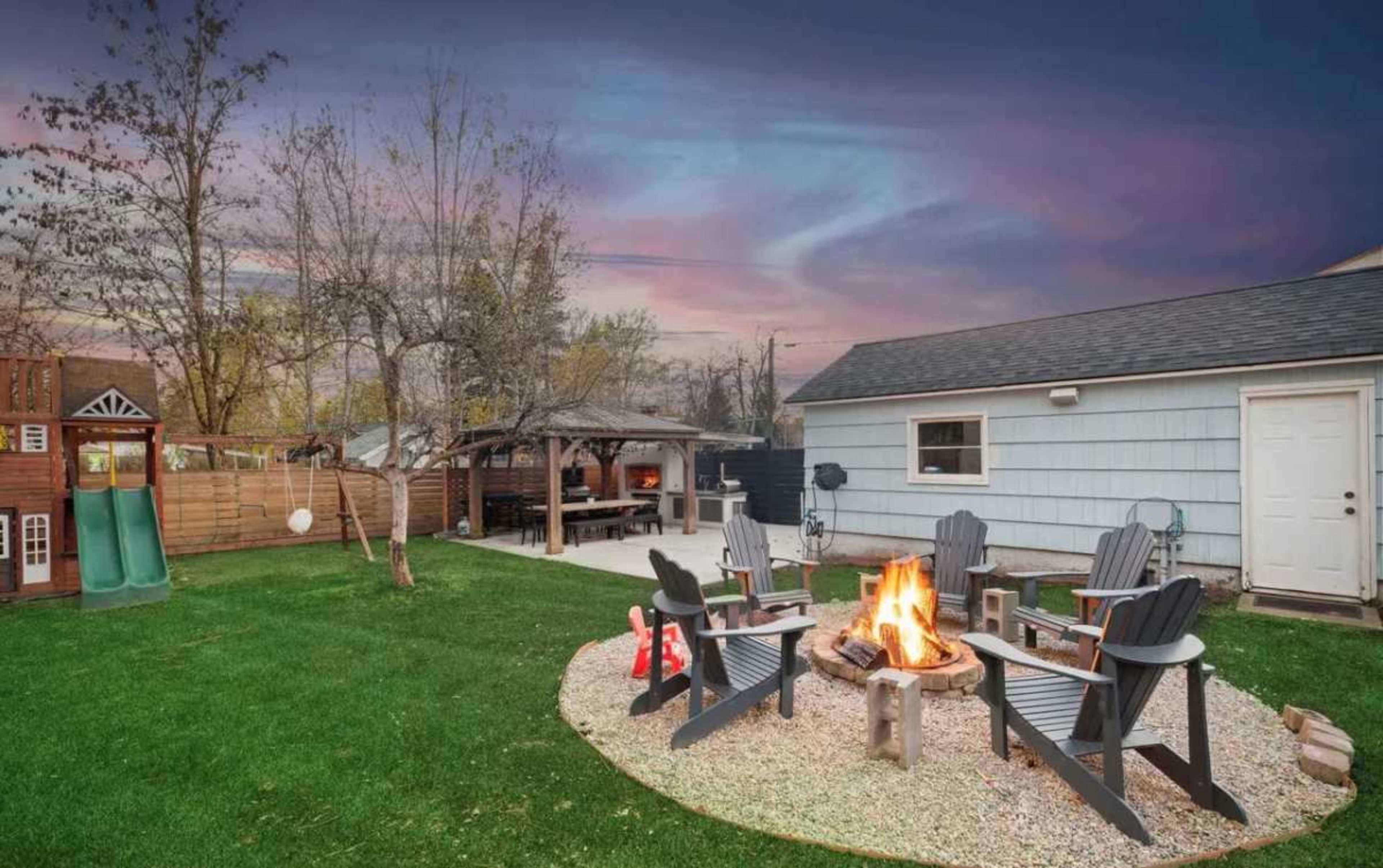 A circular fire pit surrounded by Adirondack chairs is set in a backyard with a playground and a shed, under a colorful evening sky.