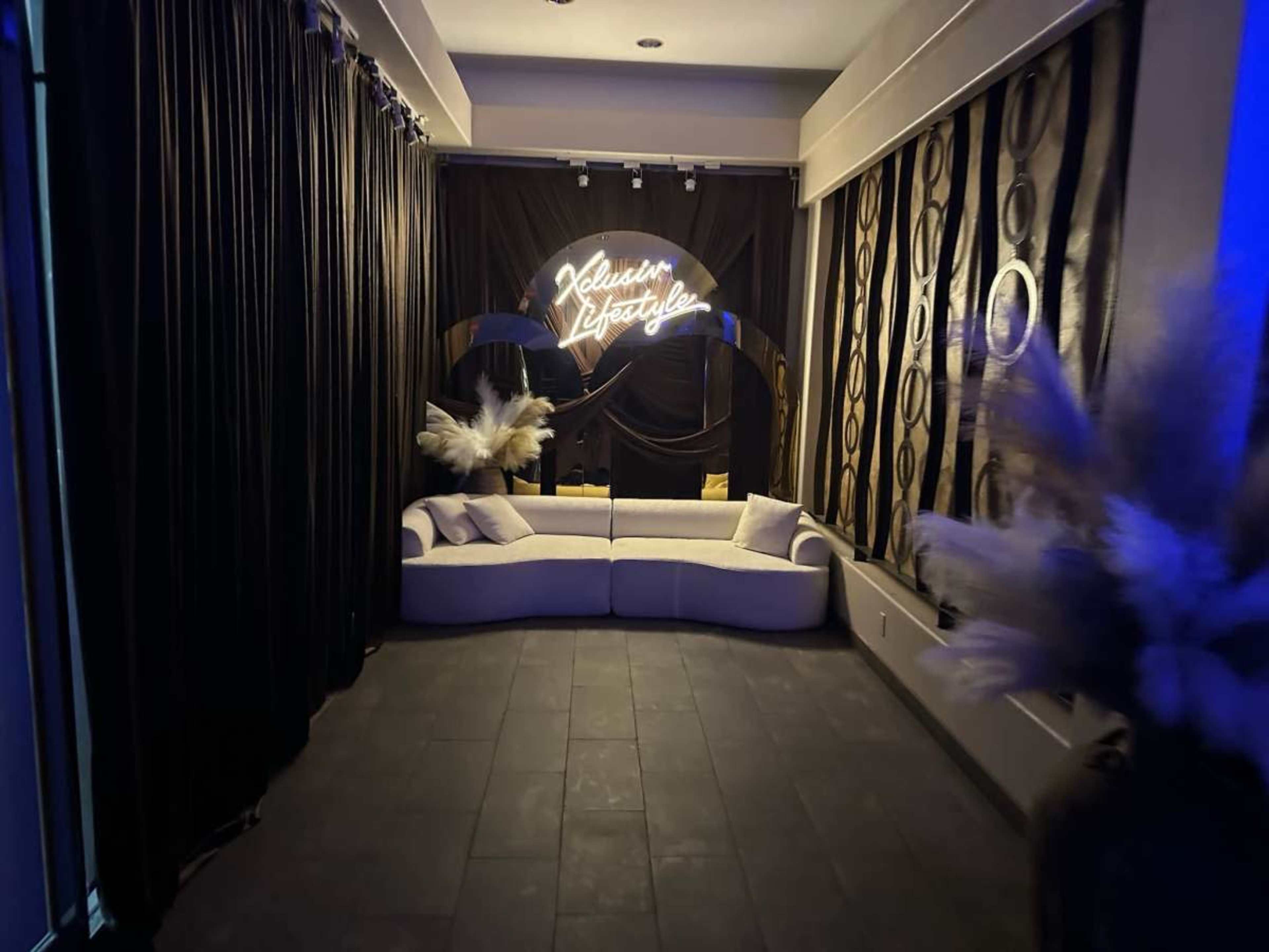 The image shows a dimly lit room with a stylish gray sofa, black curtains, and a neon sign that reads "Exclusive Lifestyle."