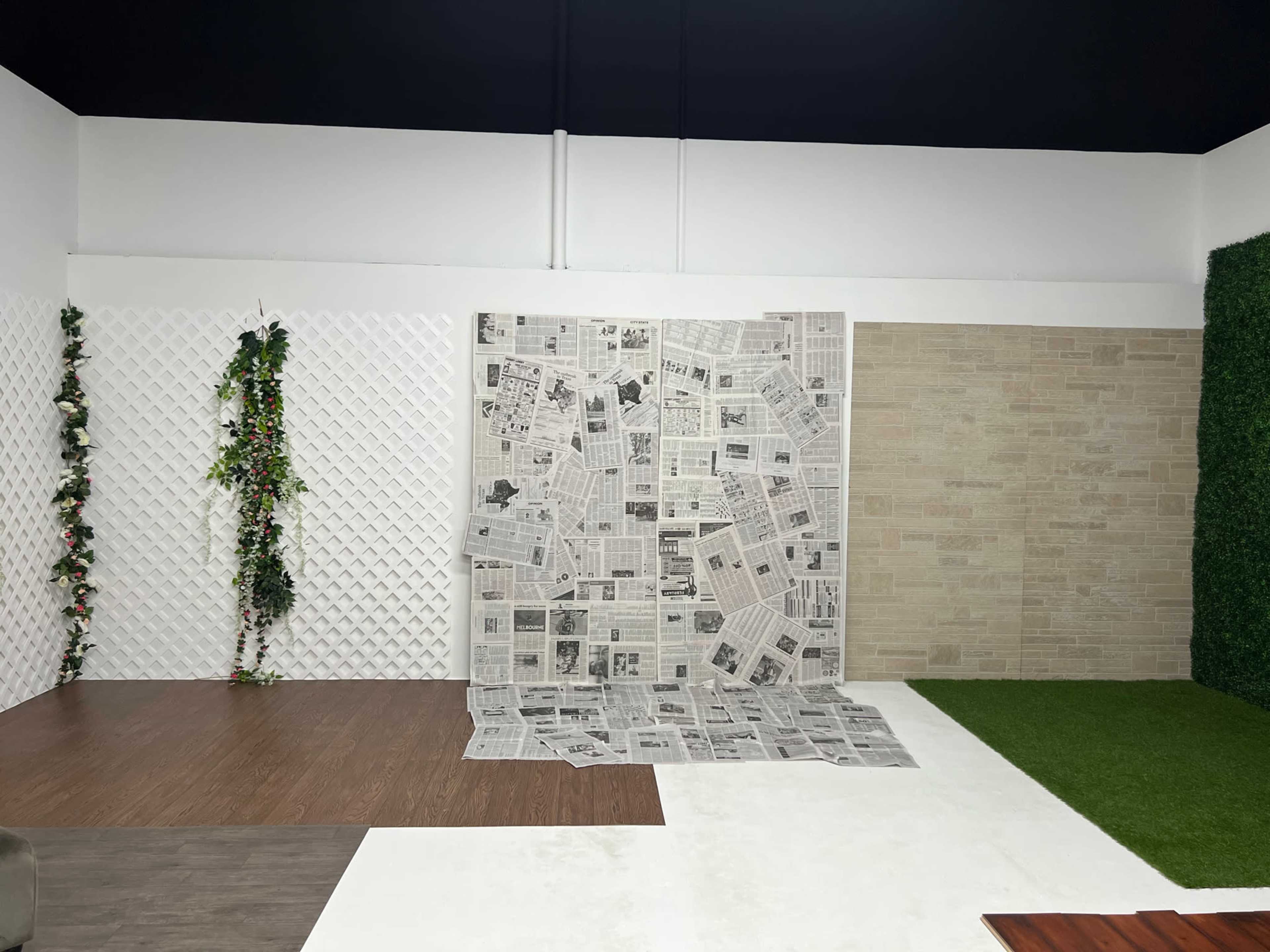 The image shows a studio space with three distinct wall textures, including a newspaper collage, a textured beige wall, and a section with artificial grass, bordered by a white lattice with hanging greenery.
