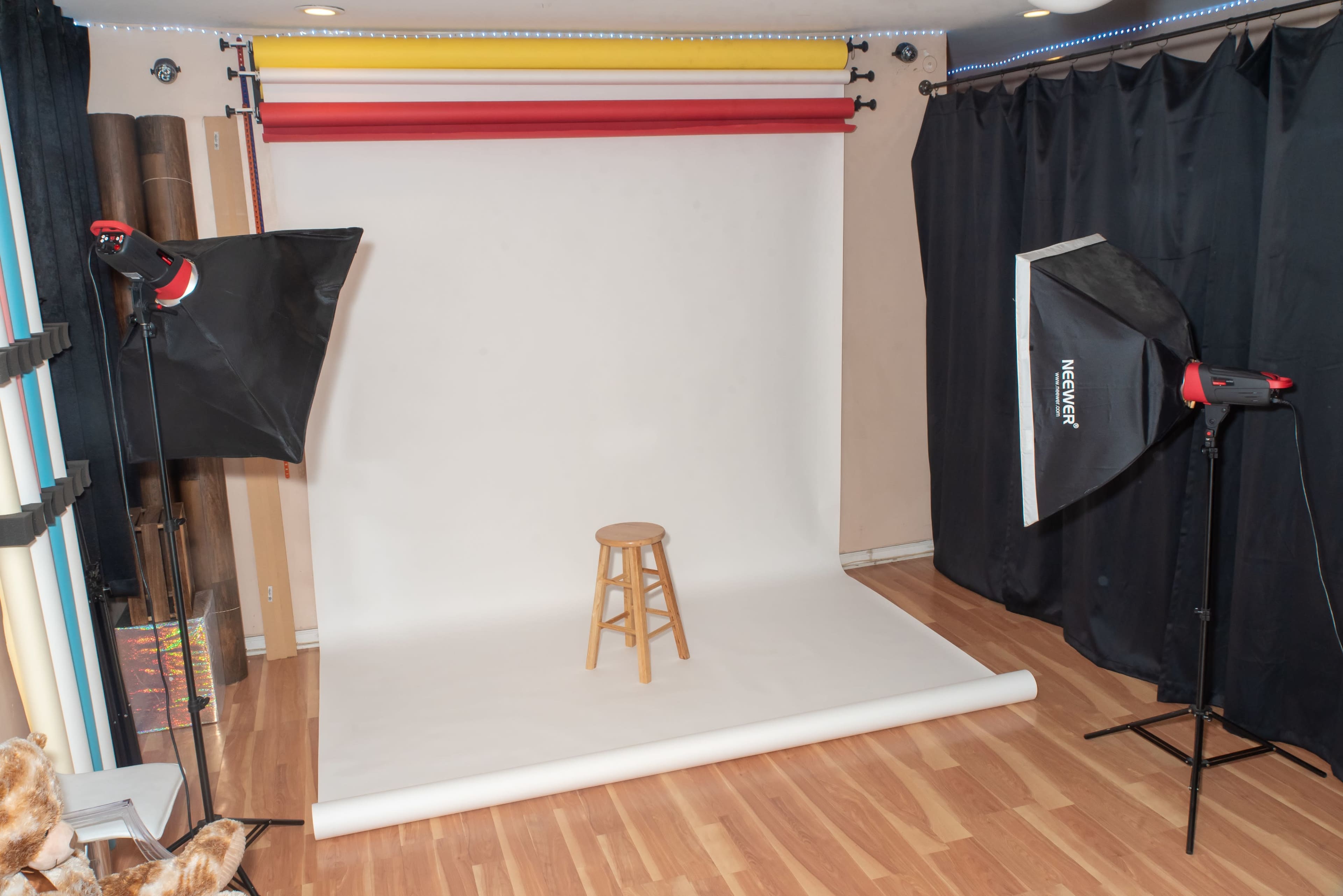 The image shows a photography studio setup featuring a white backdrop, a wooden stool in the center, and two softbox lights on either side.