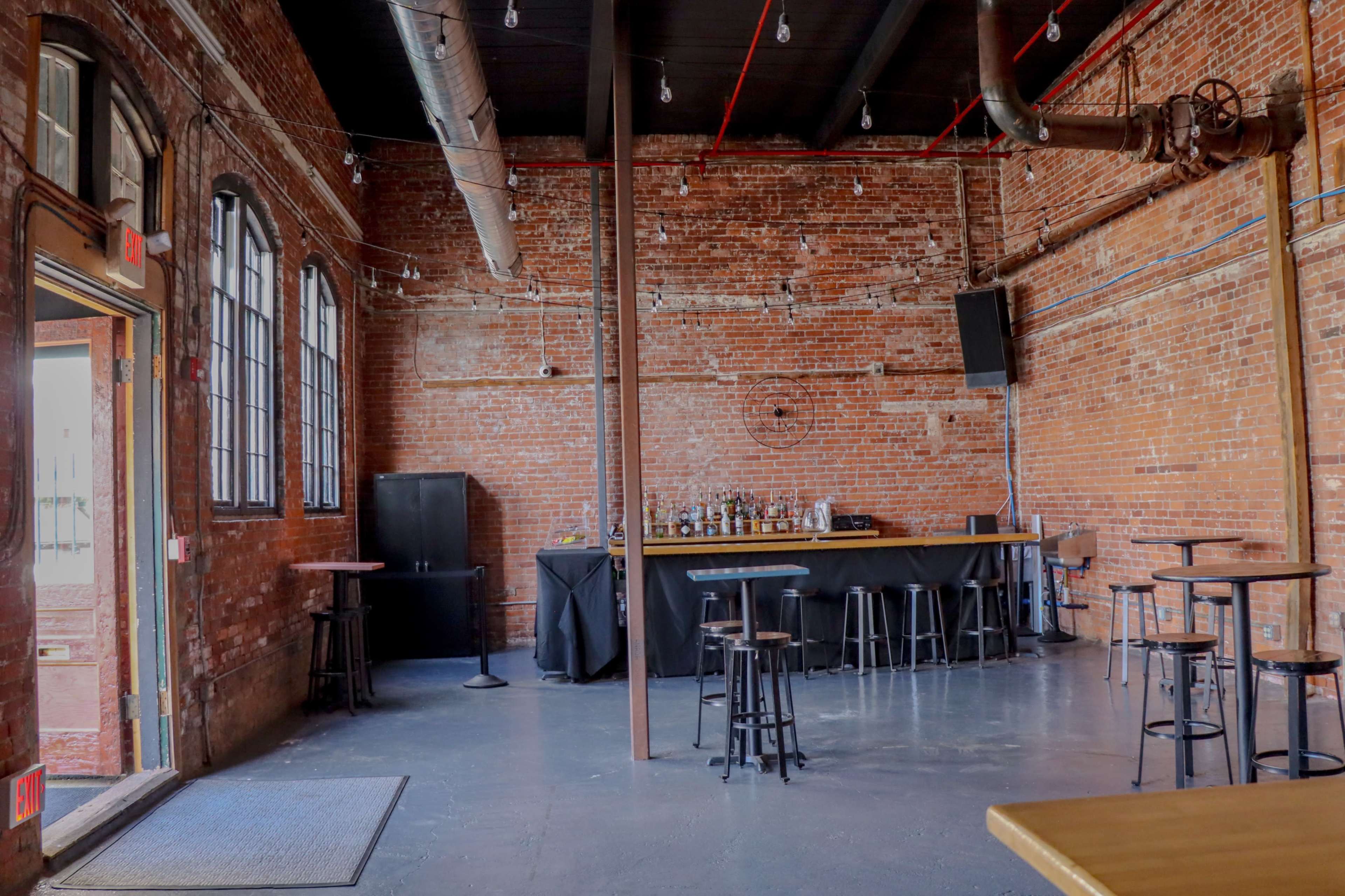 The image shows an interior space of a bar with exposed brick walls, a bar counter, and several tall tables and stools, with an open door leading outside.