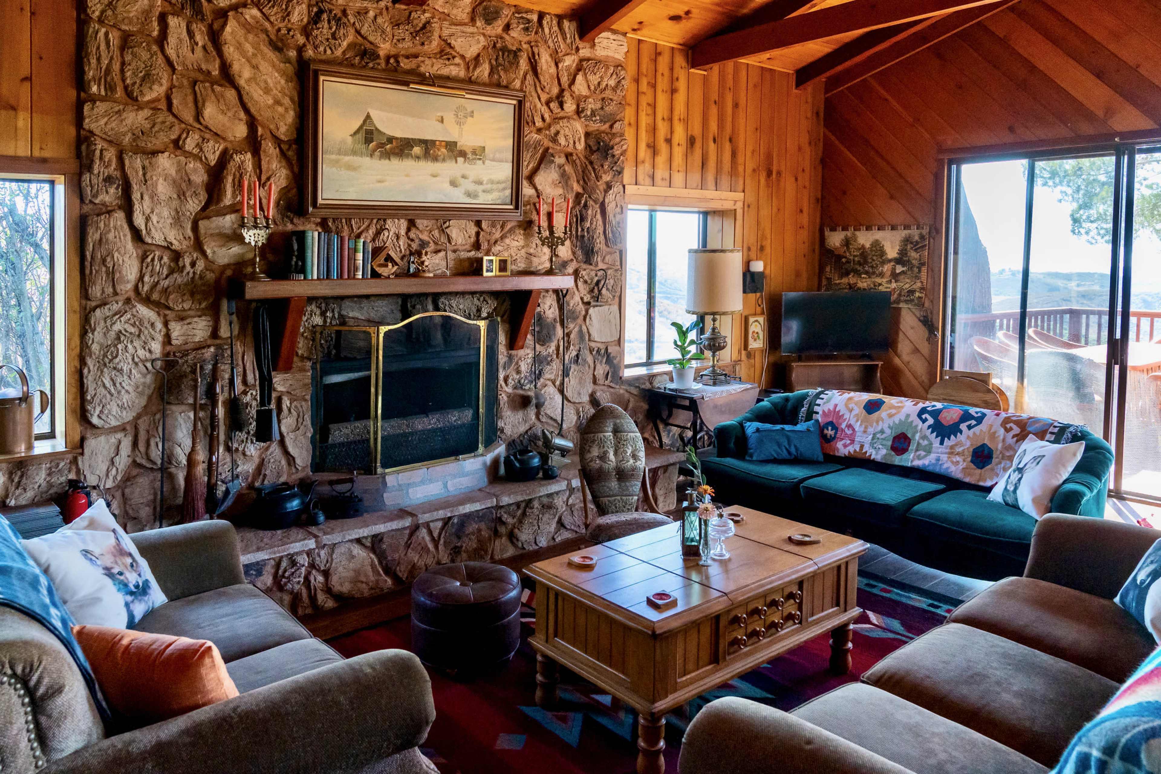 The image shows a cozy living room with stone walls, wooden beams, two sofas, a coffee table, and large windows overlooking a scenic view.