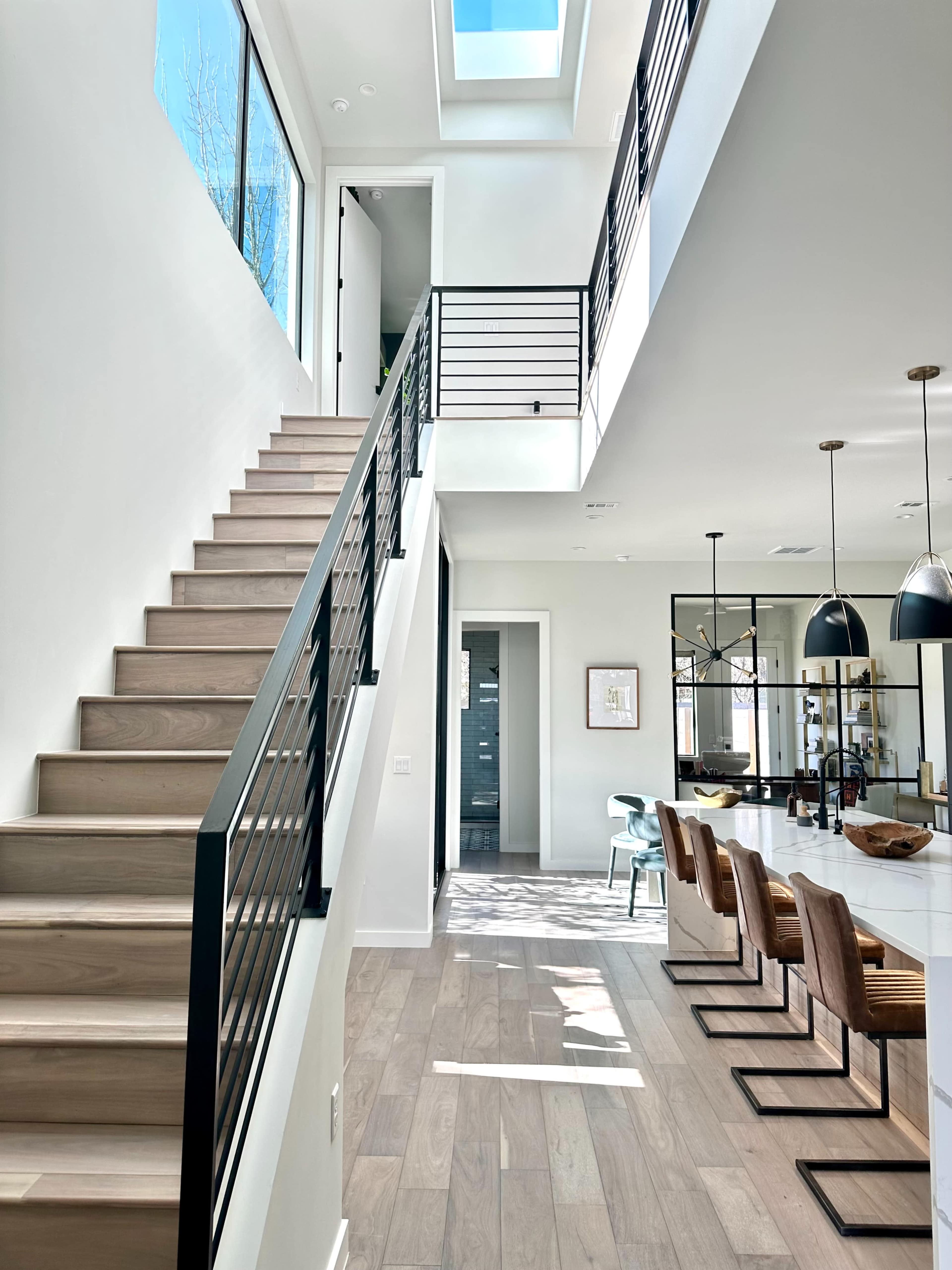 A staircase with sleek railings leads to an upper level in a modern home, featuring an open layout with a kitchen and dining area visible in the background.