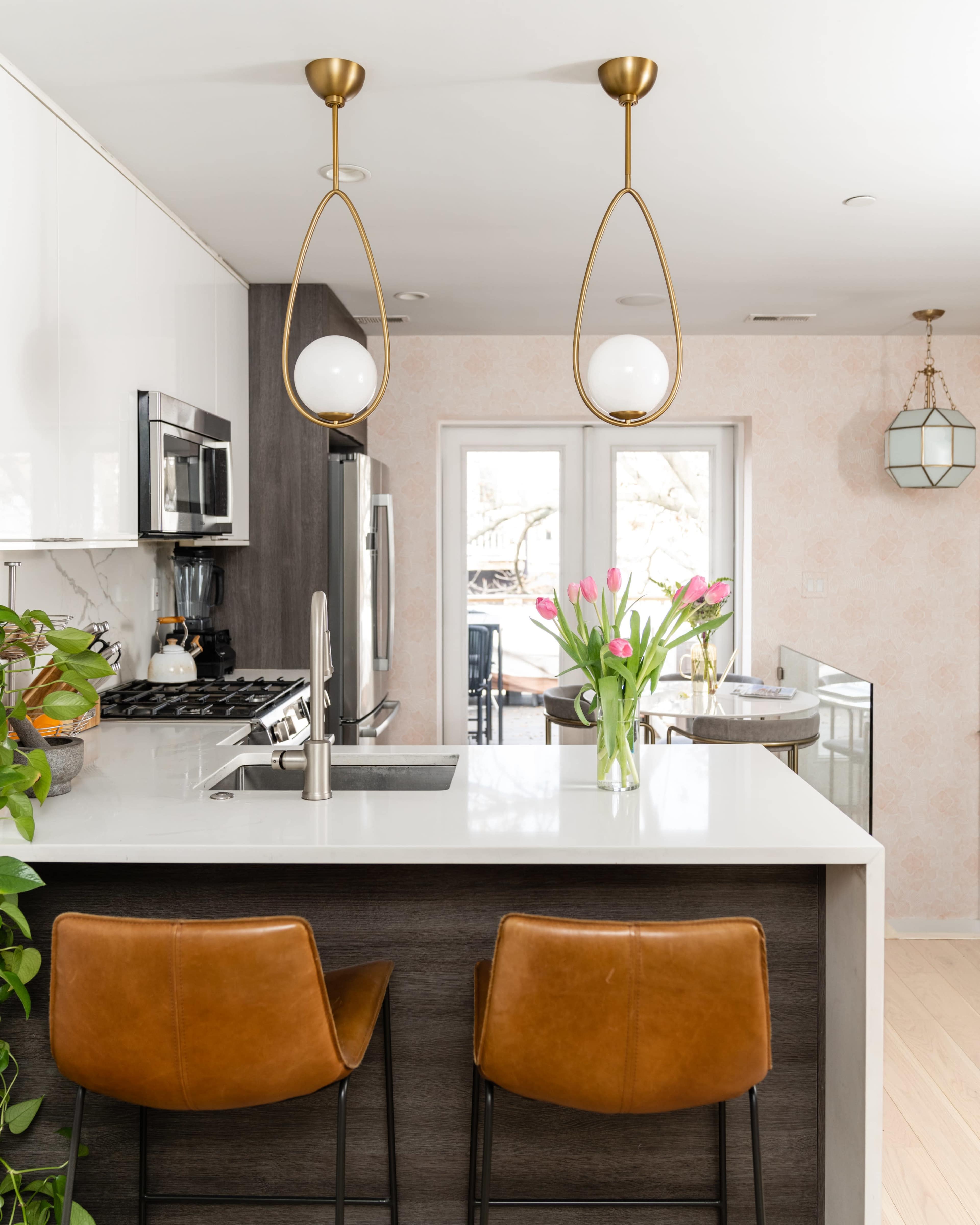 A modern kitchen features a marble island with two brown leather stools, stylish pendant lights above, and a vase of tulips on the countertop, with a view of an outdoor area beyond a glass door.