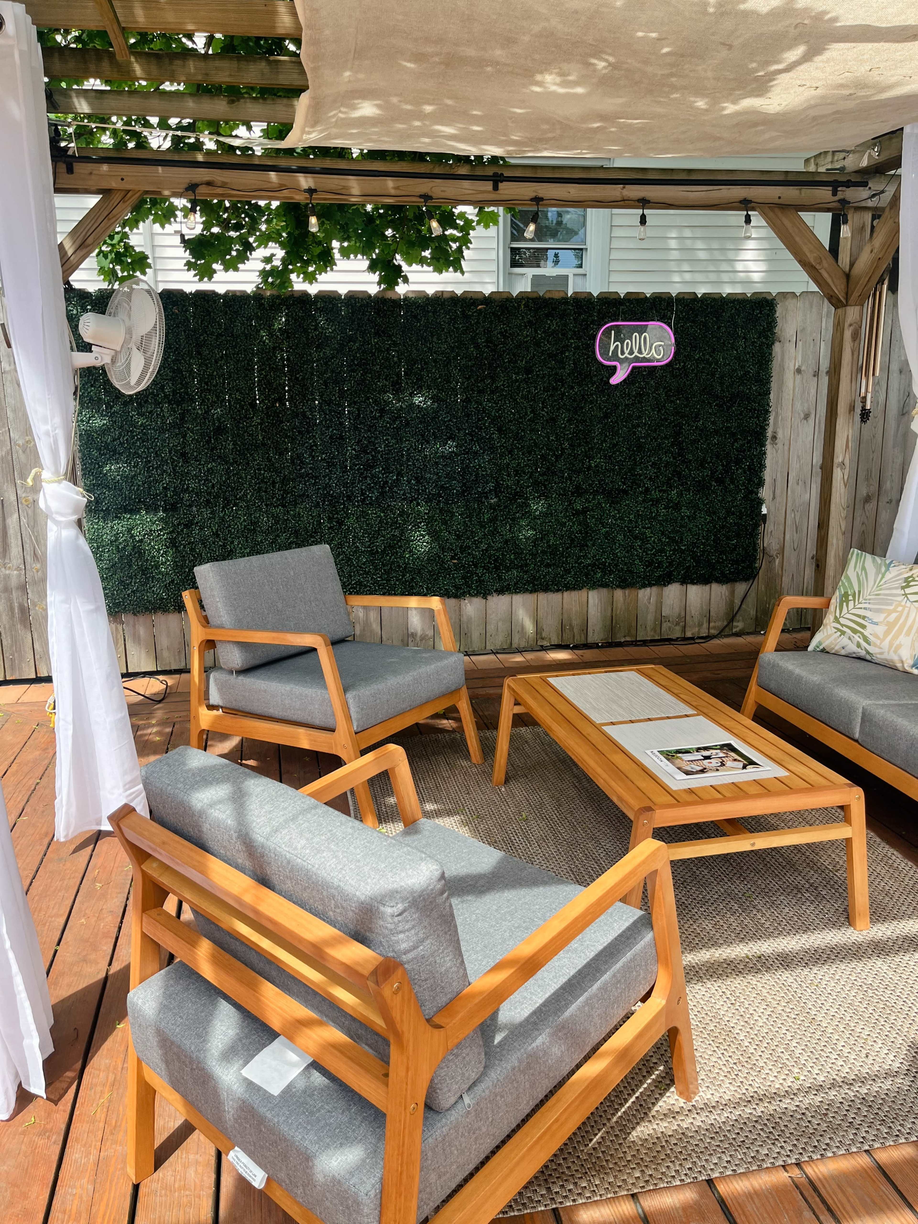 A shaded outdoor seating area features gray chairs and a wooden coffee table on a deck, surrounded by a green hedge and a "hello" sign.