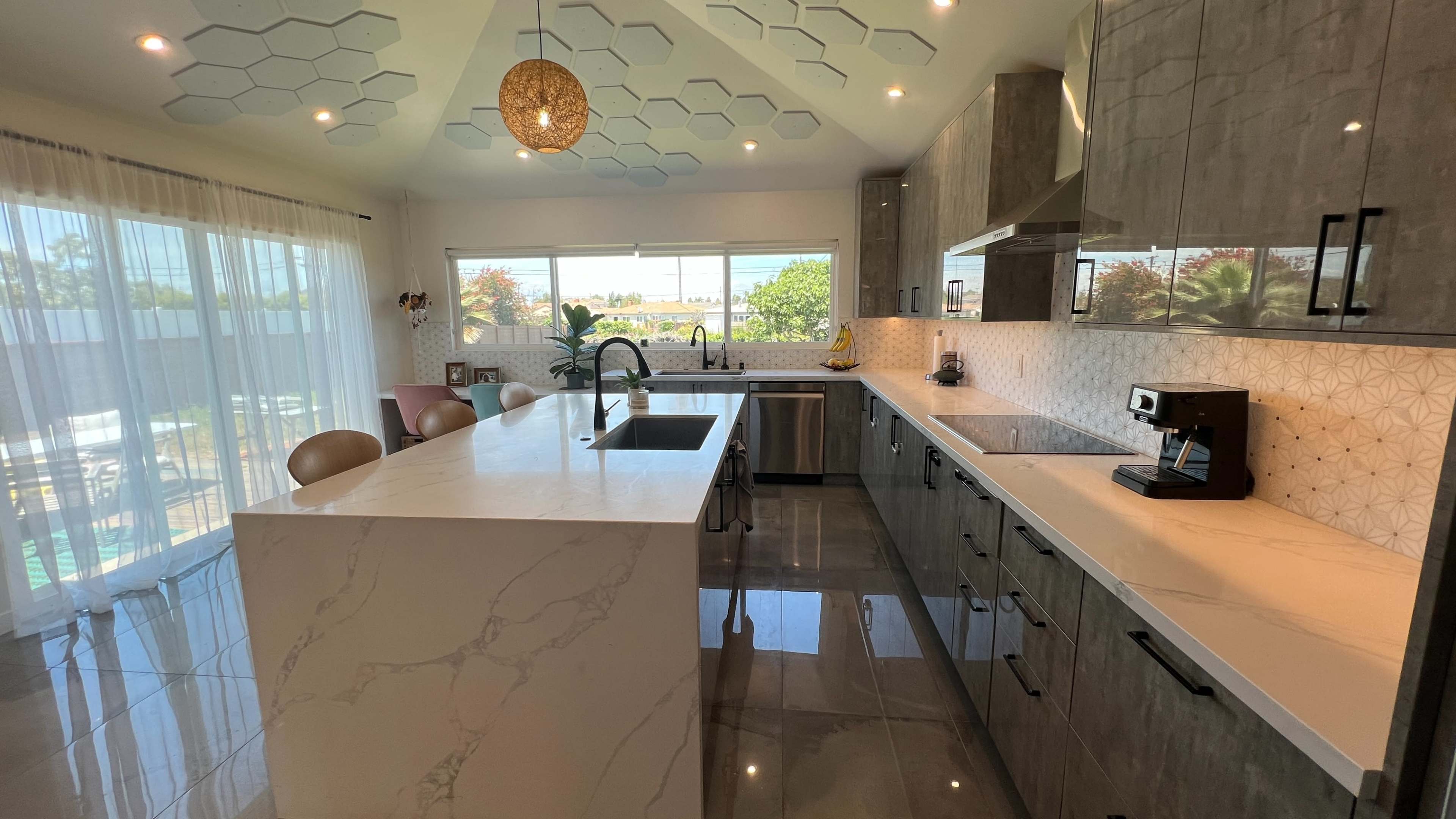 A modern kitchen with gray cabinetry, a large marble island, and a hexagonal ceiling design, illuminated by a pendant light.