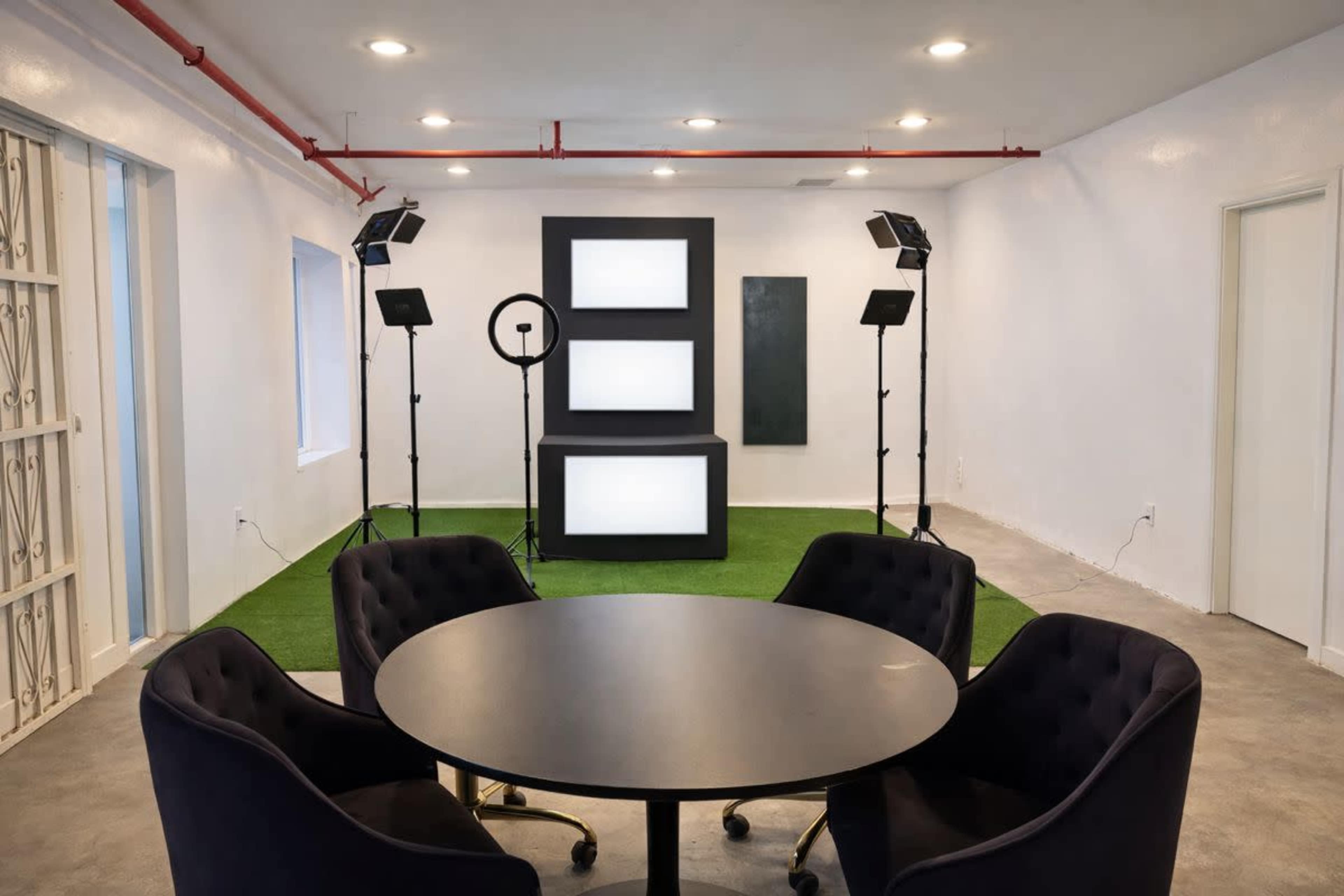 The image shows a modern room with a circular black table surrounded by four dark chairs, featuring a backdrop with illuminated panels and photography lighting equipment set up in the background.