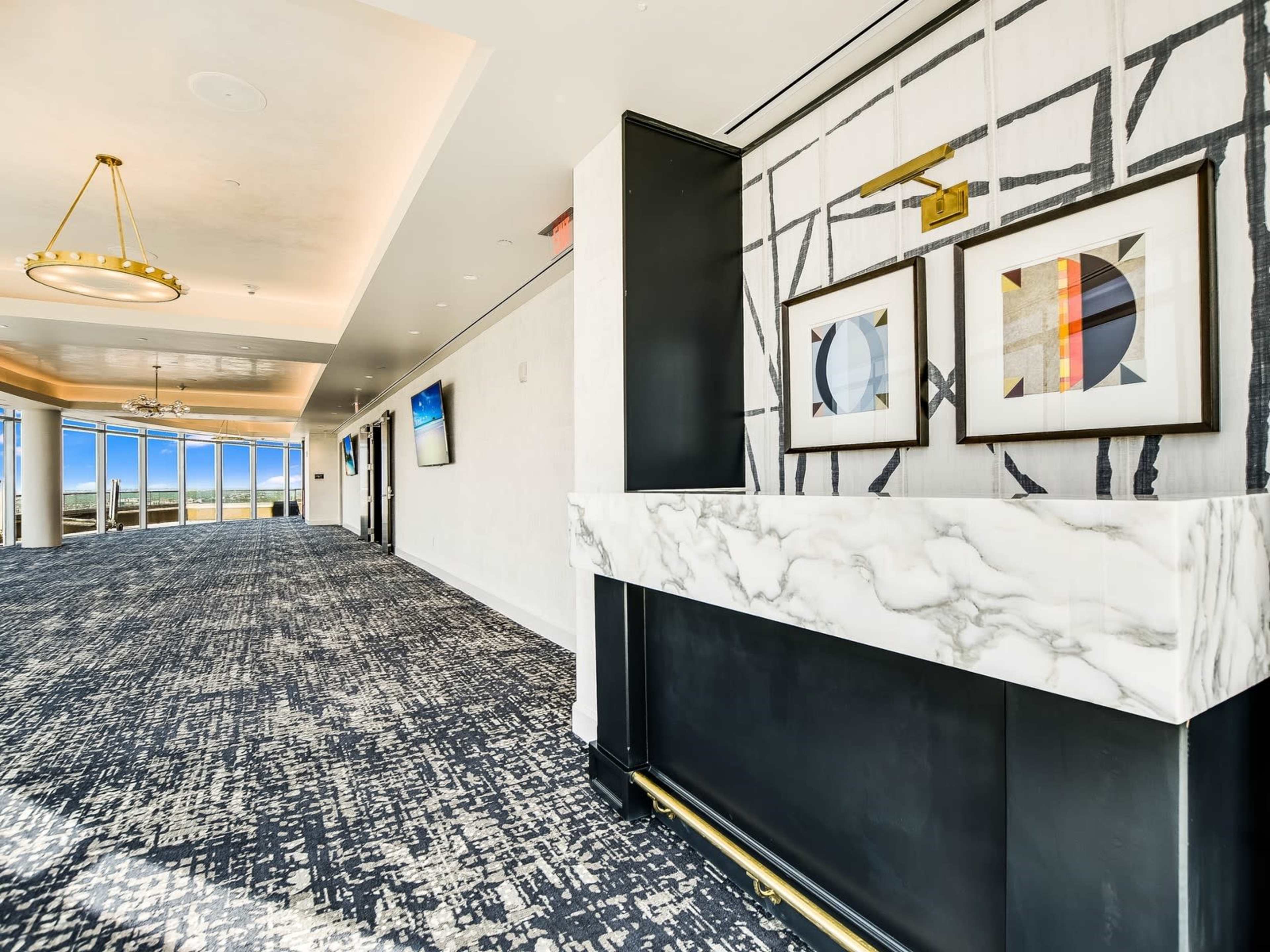 A spacious interior hallway with patterned carpeting, framed artwork on the walls, and large windows allowing natural light to enter.