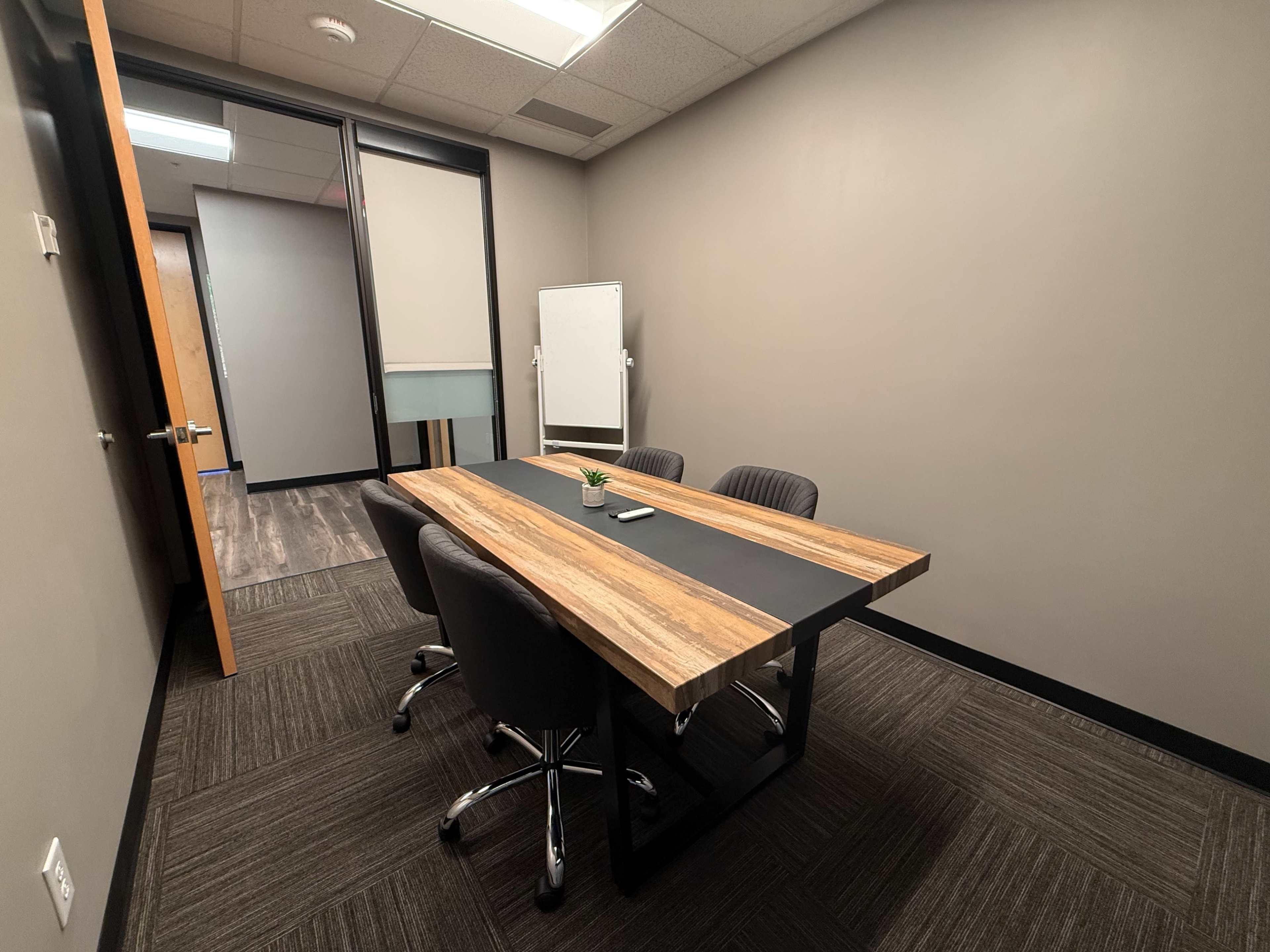 The image shows a small meeting room featuring a wooden table with four black chairs and a whiteboard in the corner.