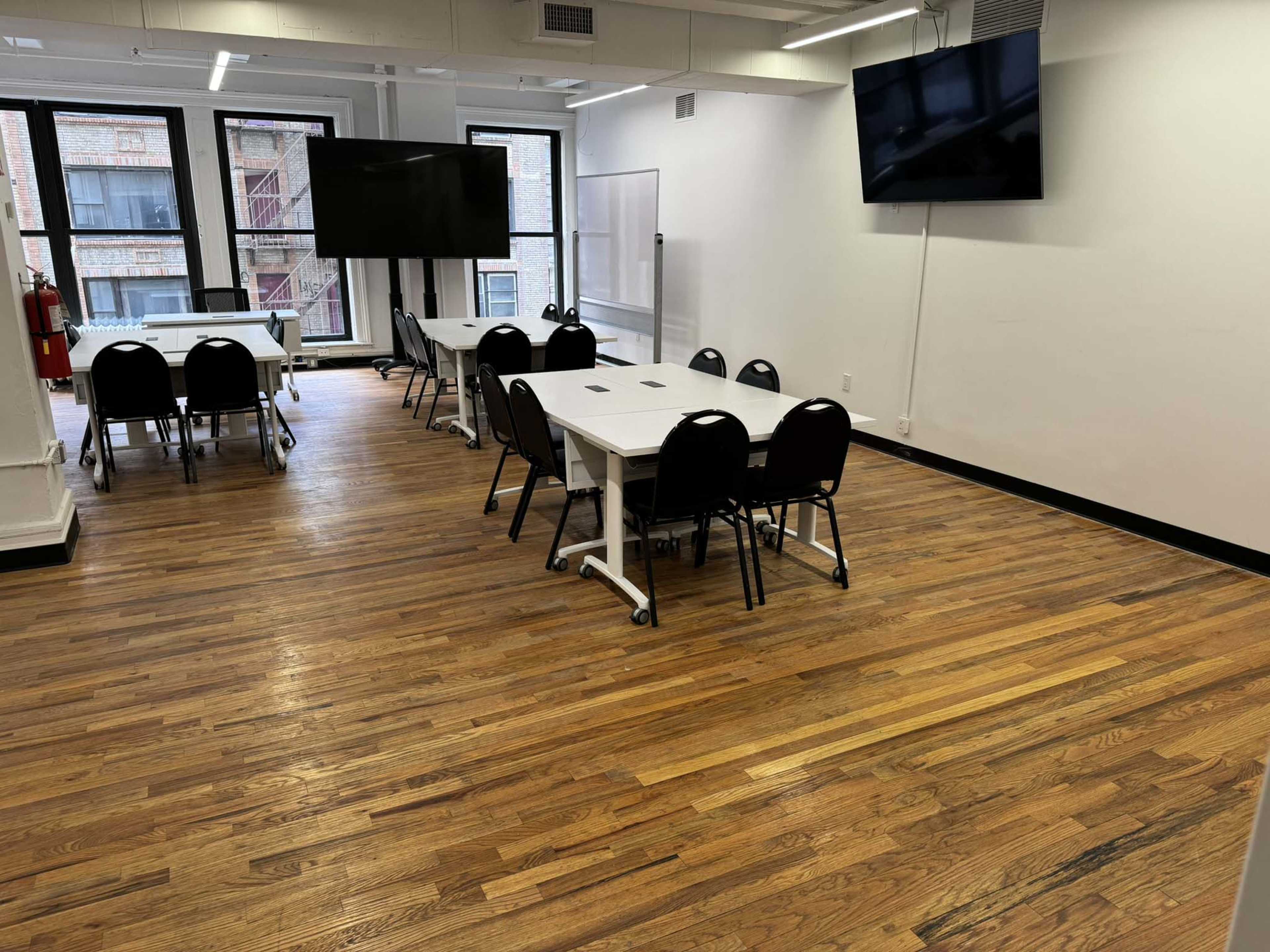 A well-lit room with wooden floors, featuring several tables and chairs arranged for a meeting or classroom setup, along with large screens mounted on the walls.