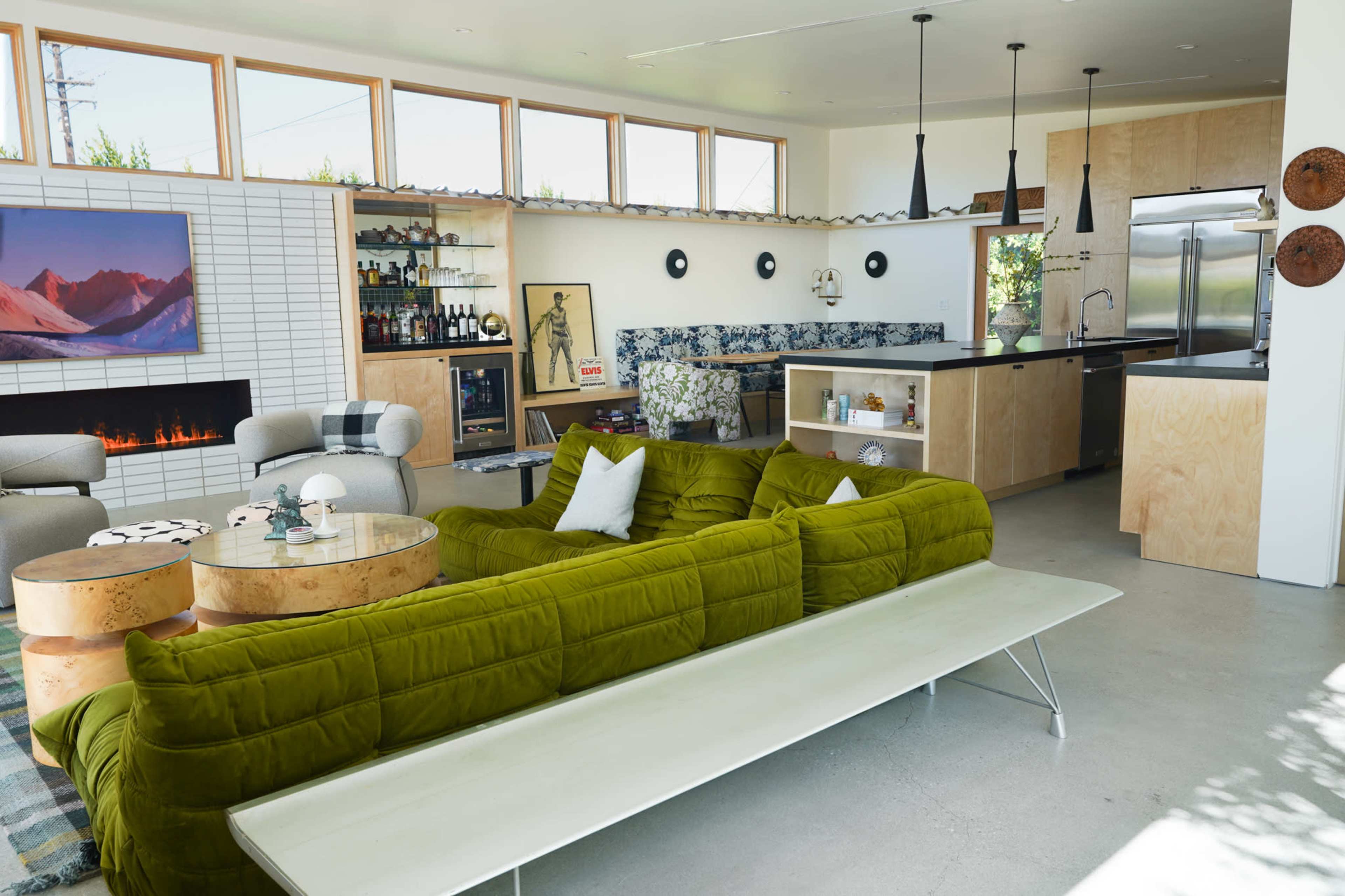 A modern living space featuring a green sectional sofa, a minimalist coffee table, a kitchen area, and a fireplace with large windows letting in natural light.