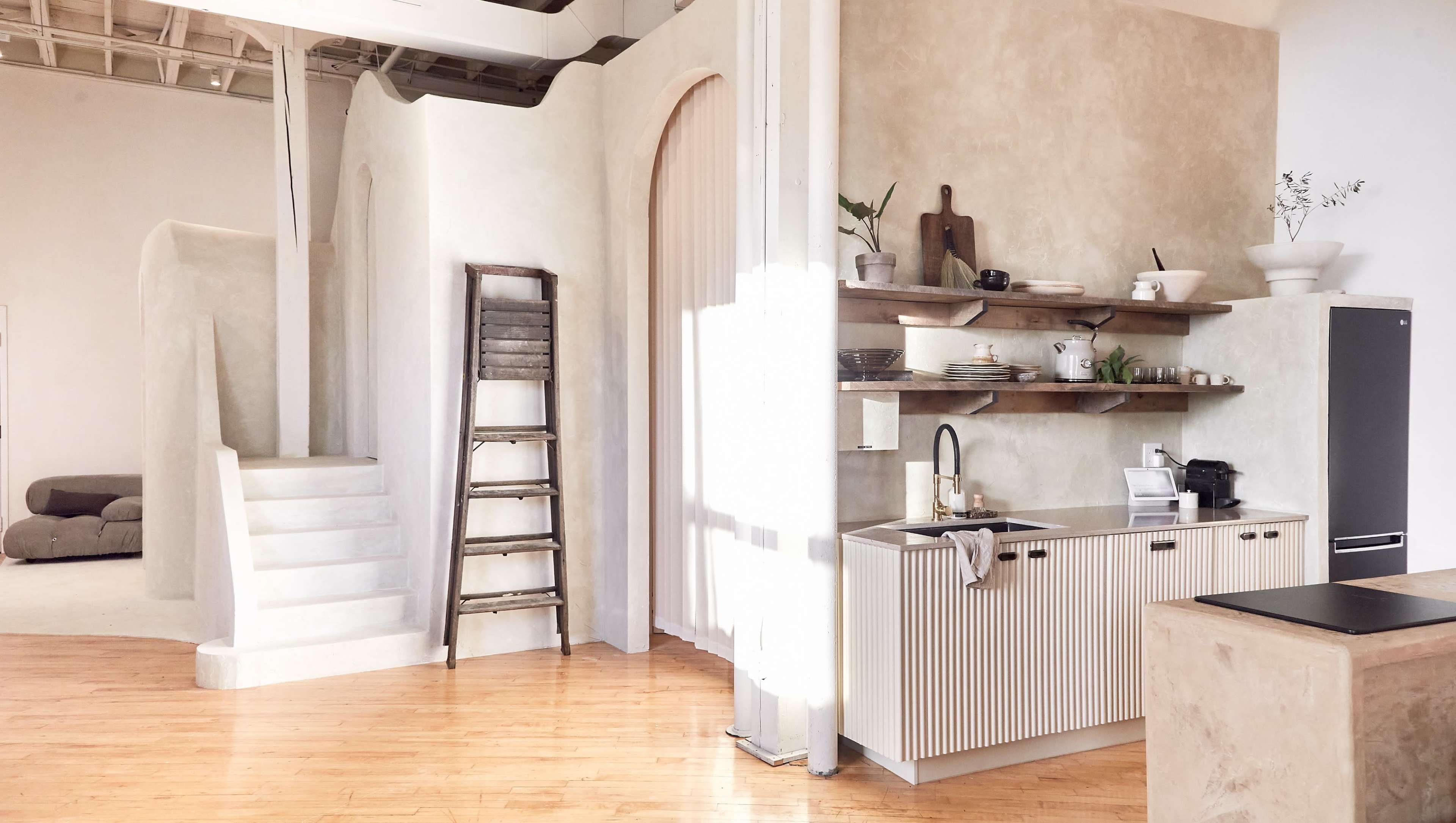 A minimalist interior space featuring a kitchen with a shelf and a ladder, alongside stairs leading to a different area with soft furniture.