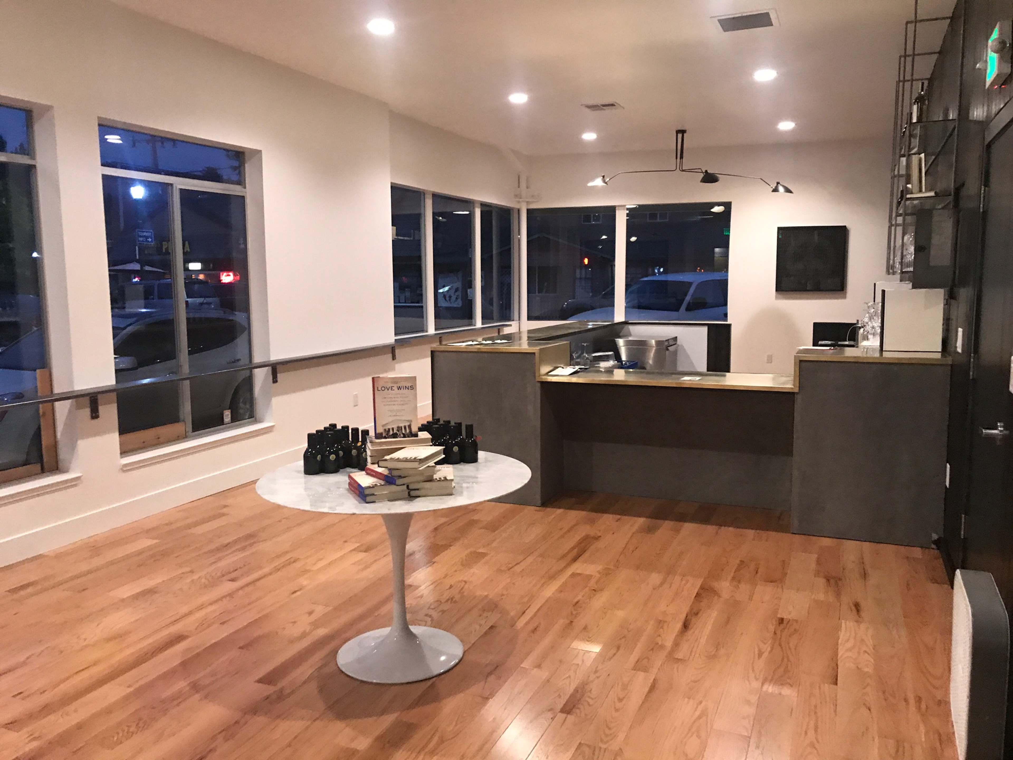 A modern, minimalistic interior space with wooden flooring, large windows, and a circular table at the center, complemented by a reception area featuring a countertop and various items.