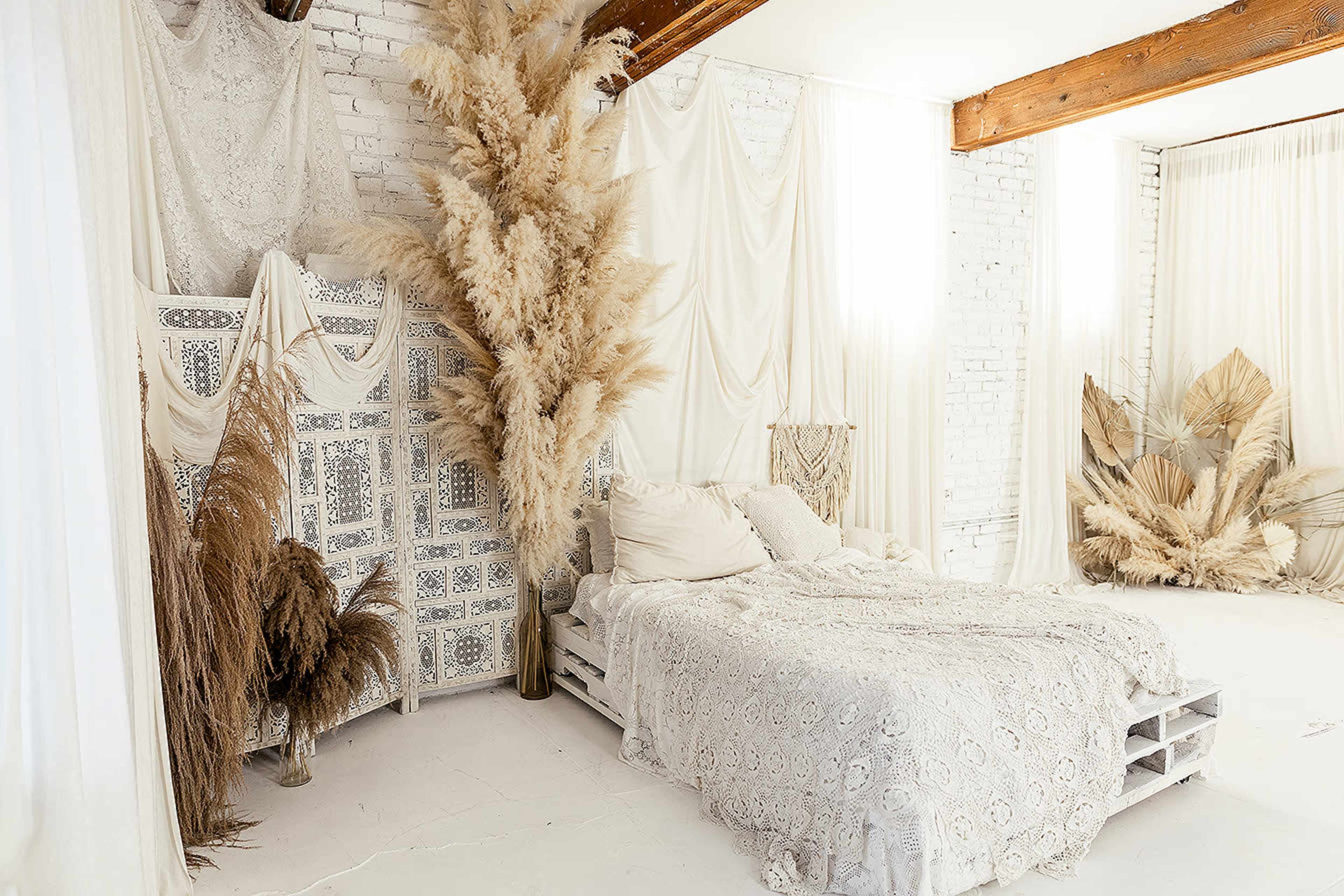 A cozy bedroom features a bed adorned with a lace coverlet, surrounded by beige plants and draped fabric against white brick walls.