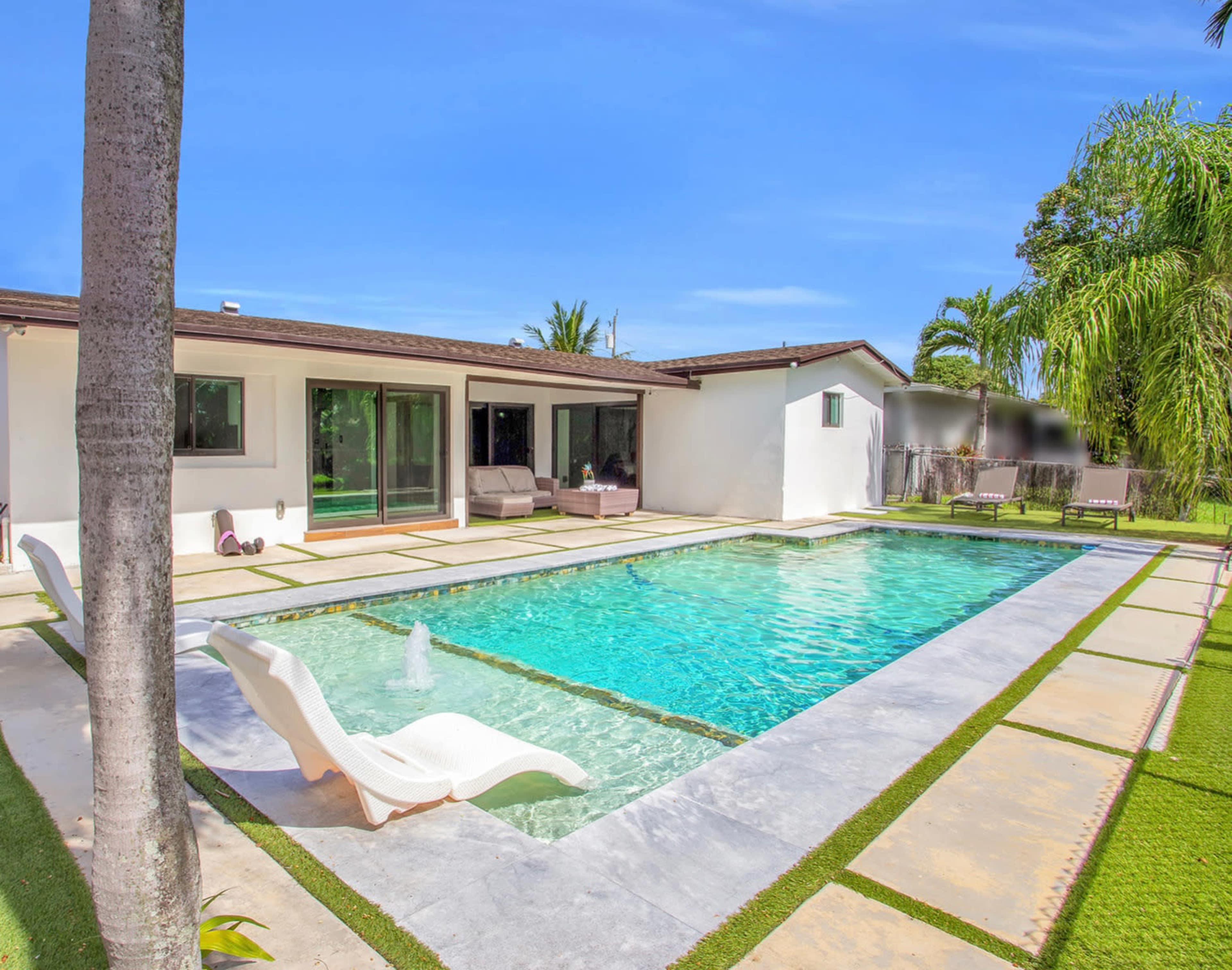 A modern, single-story house features a rectangular swimming pool surrounded by stone tiles and well-maintained grass.