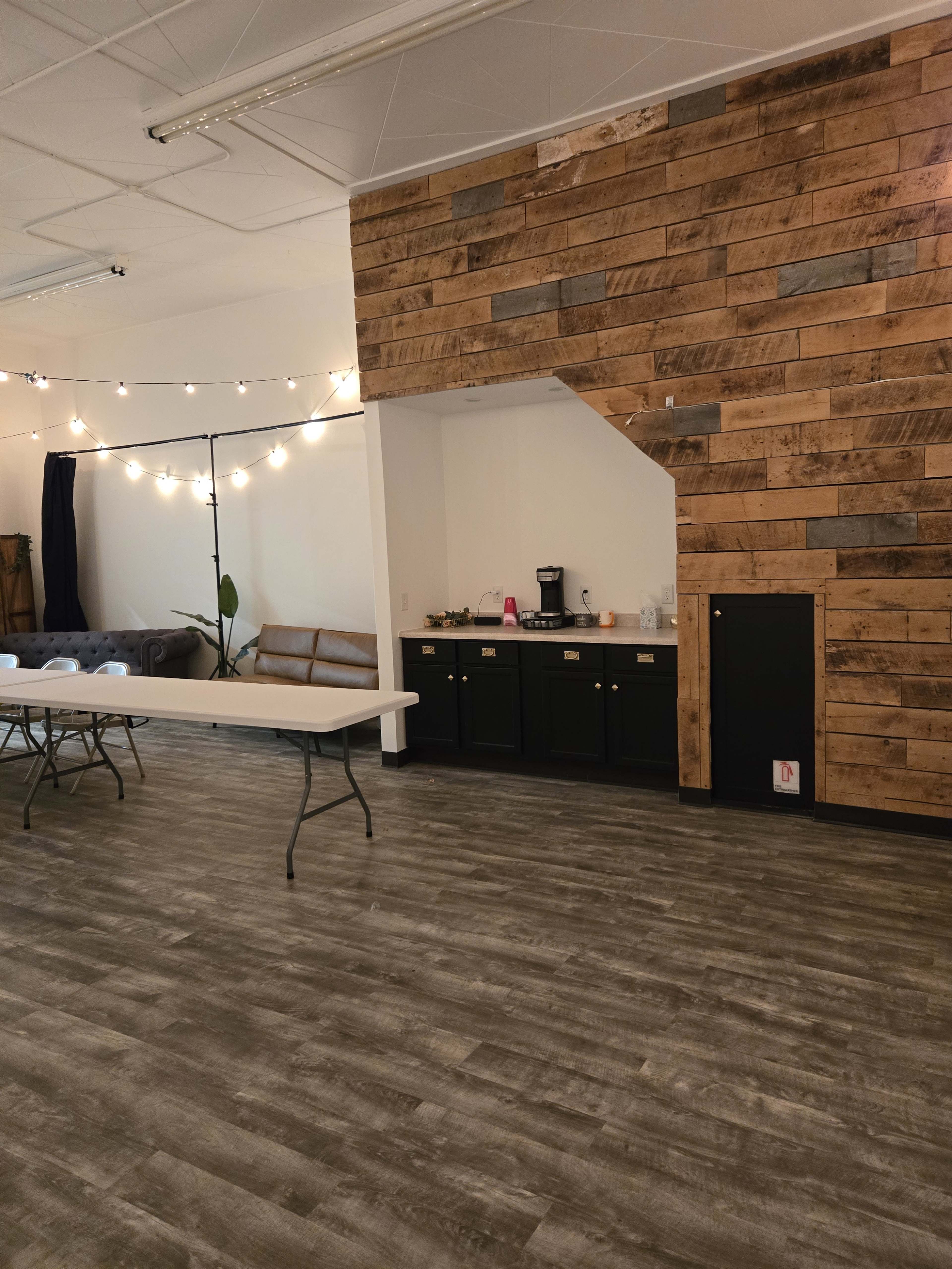 The image shows an interior room featuring a wooden accent wall, a table with folding chairs, and a beverage station against a backdrop of string lights.