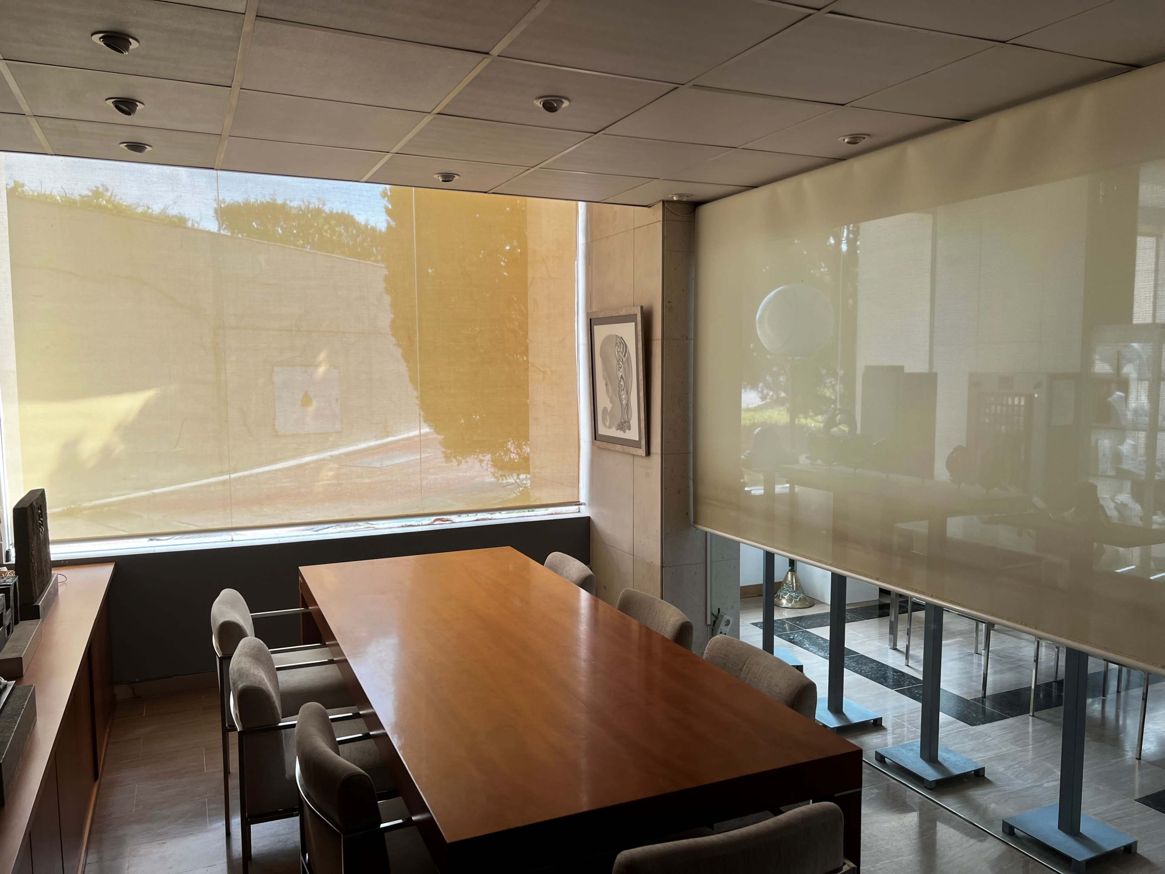The image shows a conference room with a large wooden table surrounded by chairs, featuring bright natural light filtering through sheer curtains.