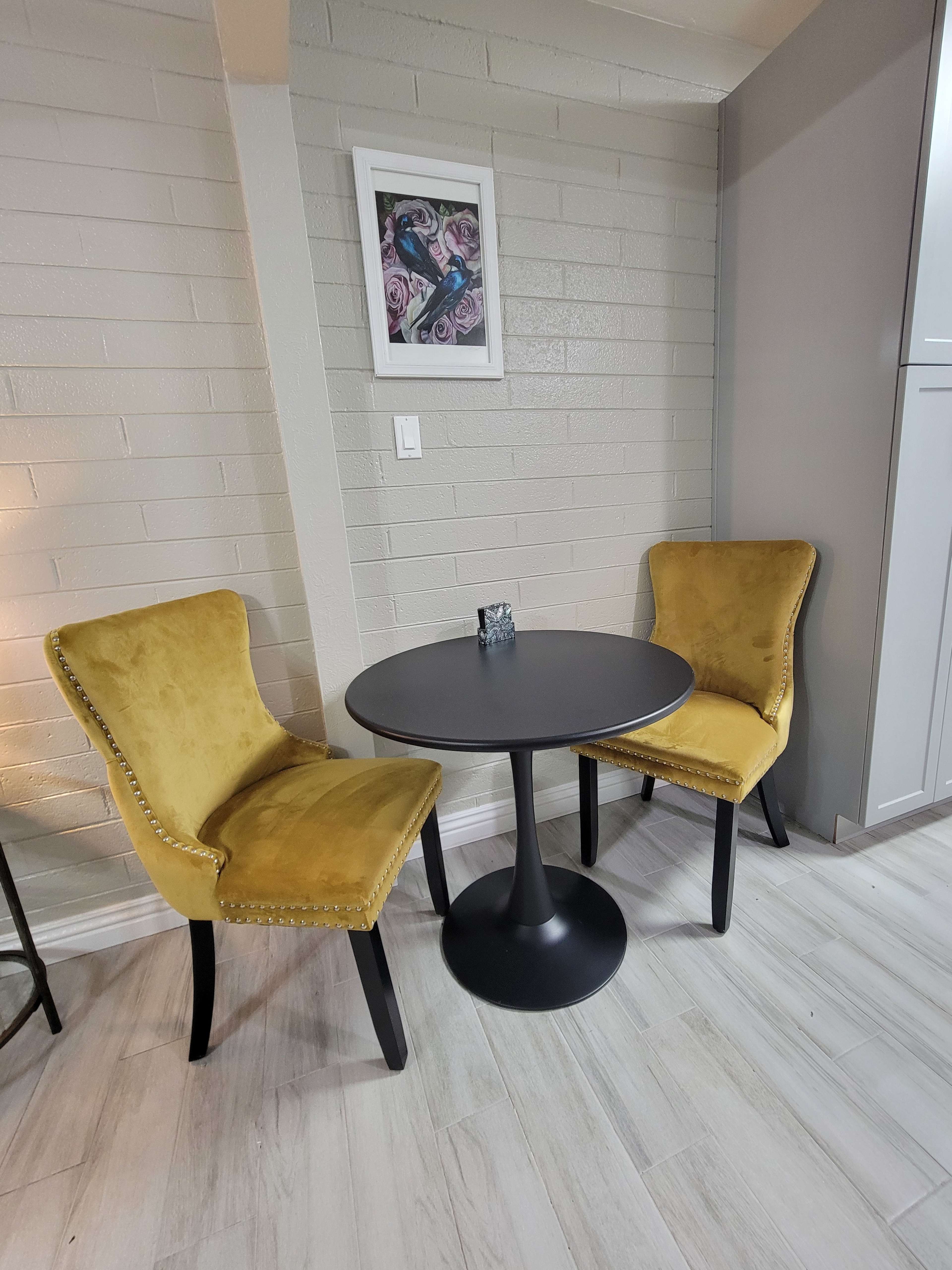 A round black table is positioned between two yellow upholstered chairs against a light gray brick wall.