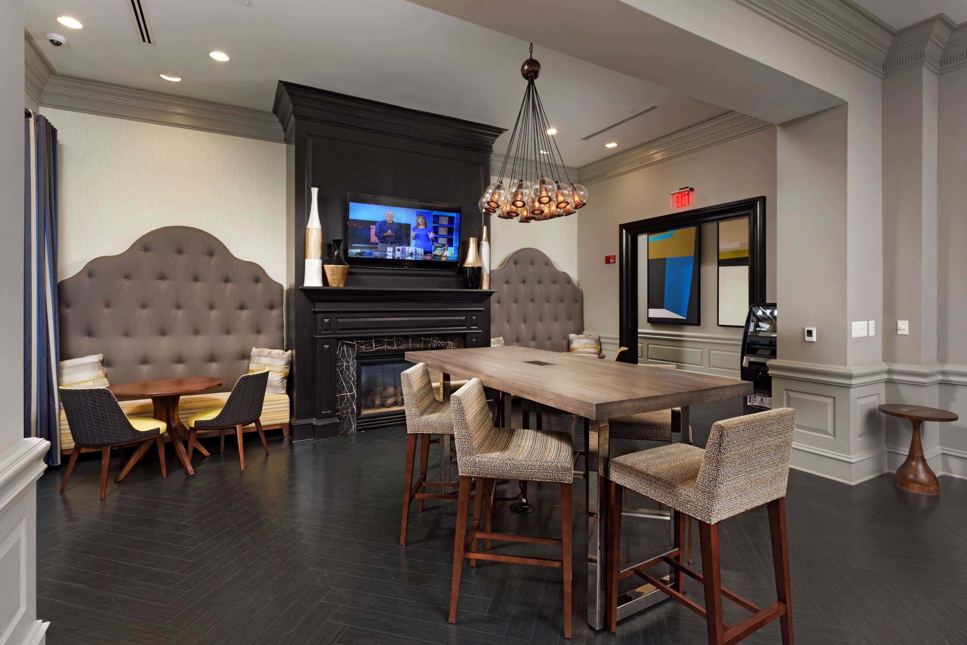 A modern lounge area featuring a large table with tall chairs, cozy seating nooks, a fireplace, and a TV mounted above the mantel.