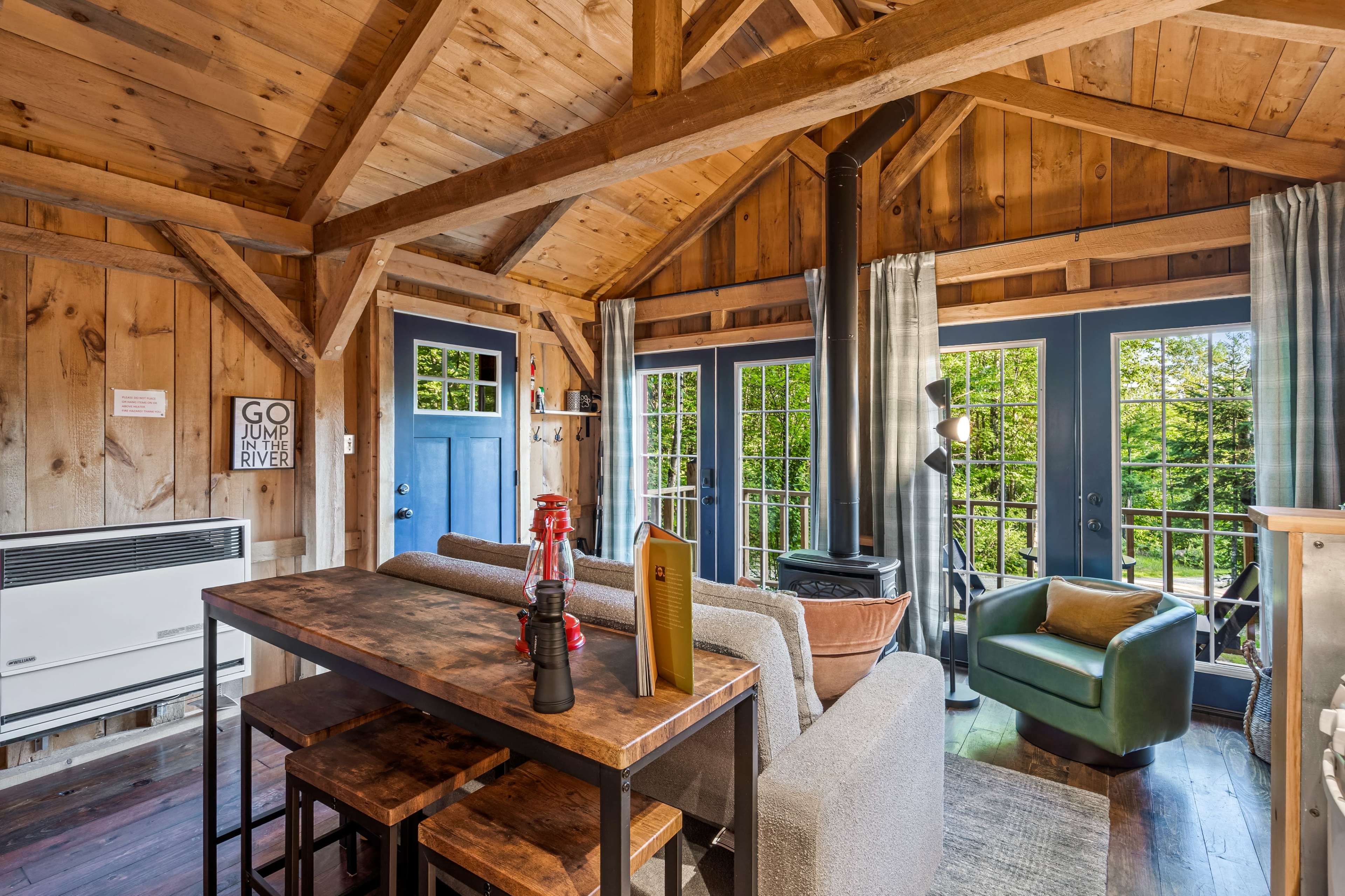 The image shows a cozy cabin interior with wooden beams, a dining table, a sofa, a green armchair, and large windows opening to a view of greenery outside.