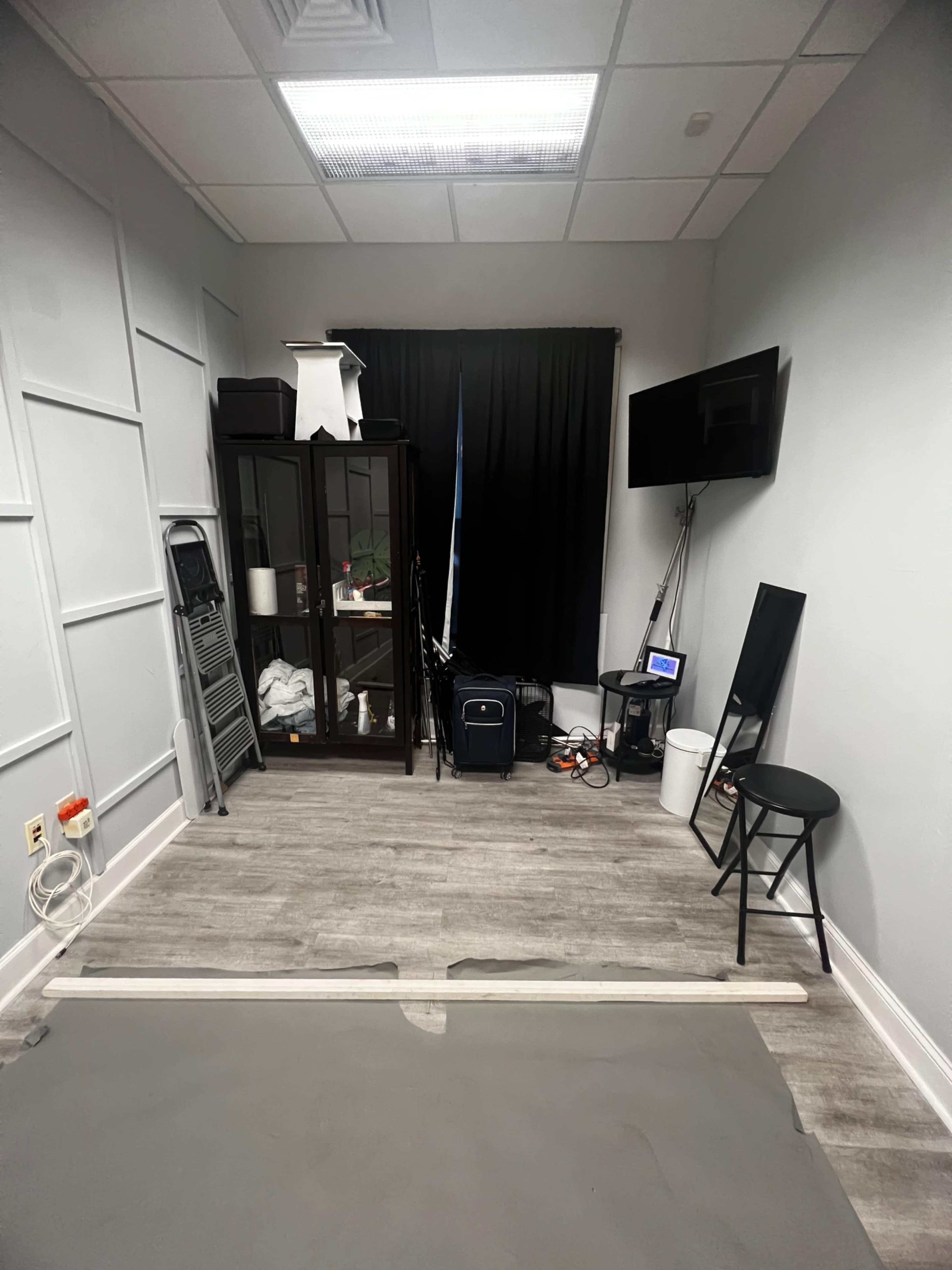 A small, sparsely furnished room with a black curtain, a display cabinet, a television, a stool, and various equipment along the walls.