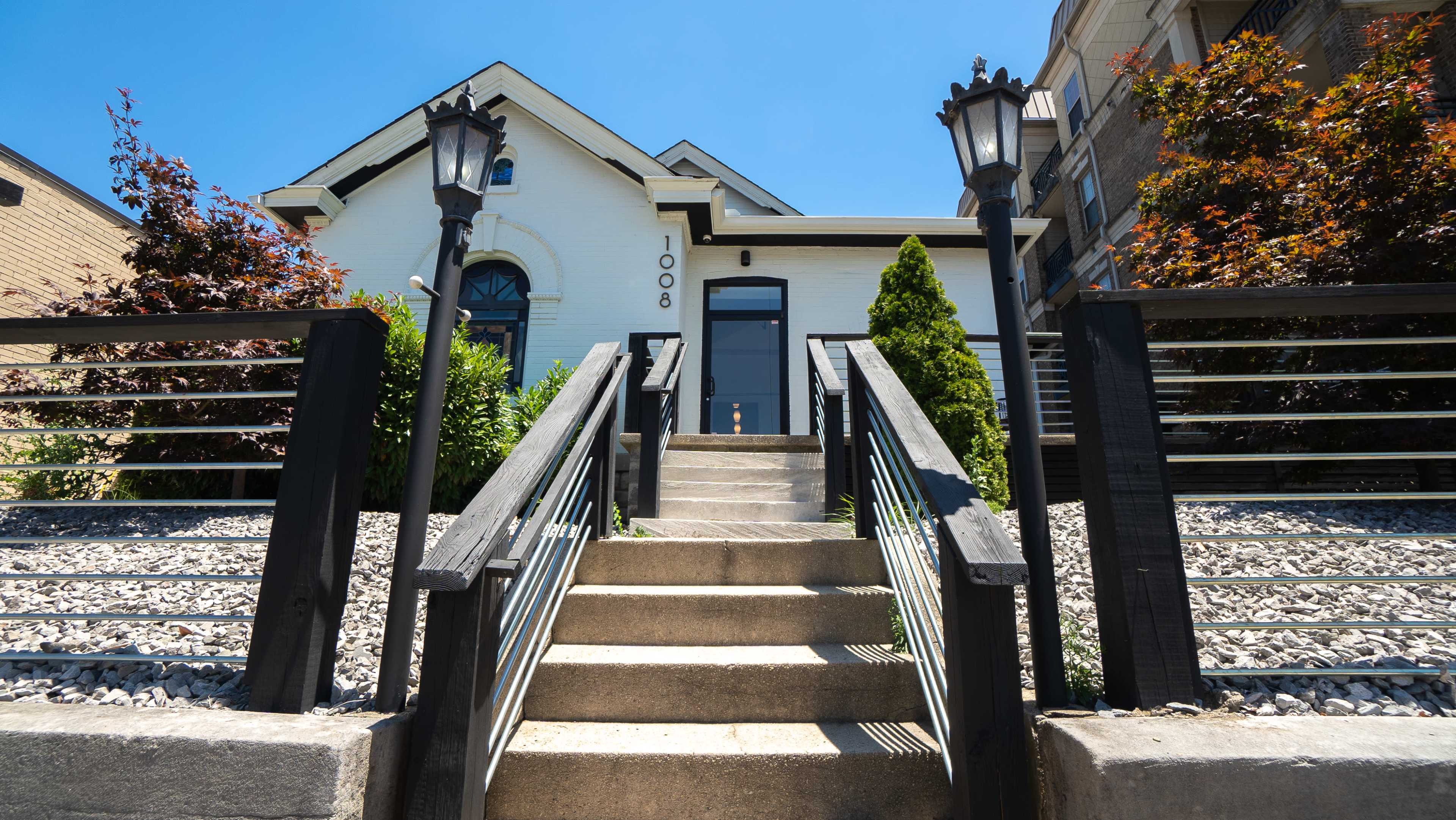 A set of concrete steps leads up to a white house with a black door, flanked by lampposts and surrounded by neatly arranged gravel and shrubs.