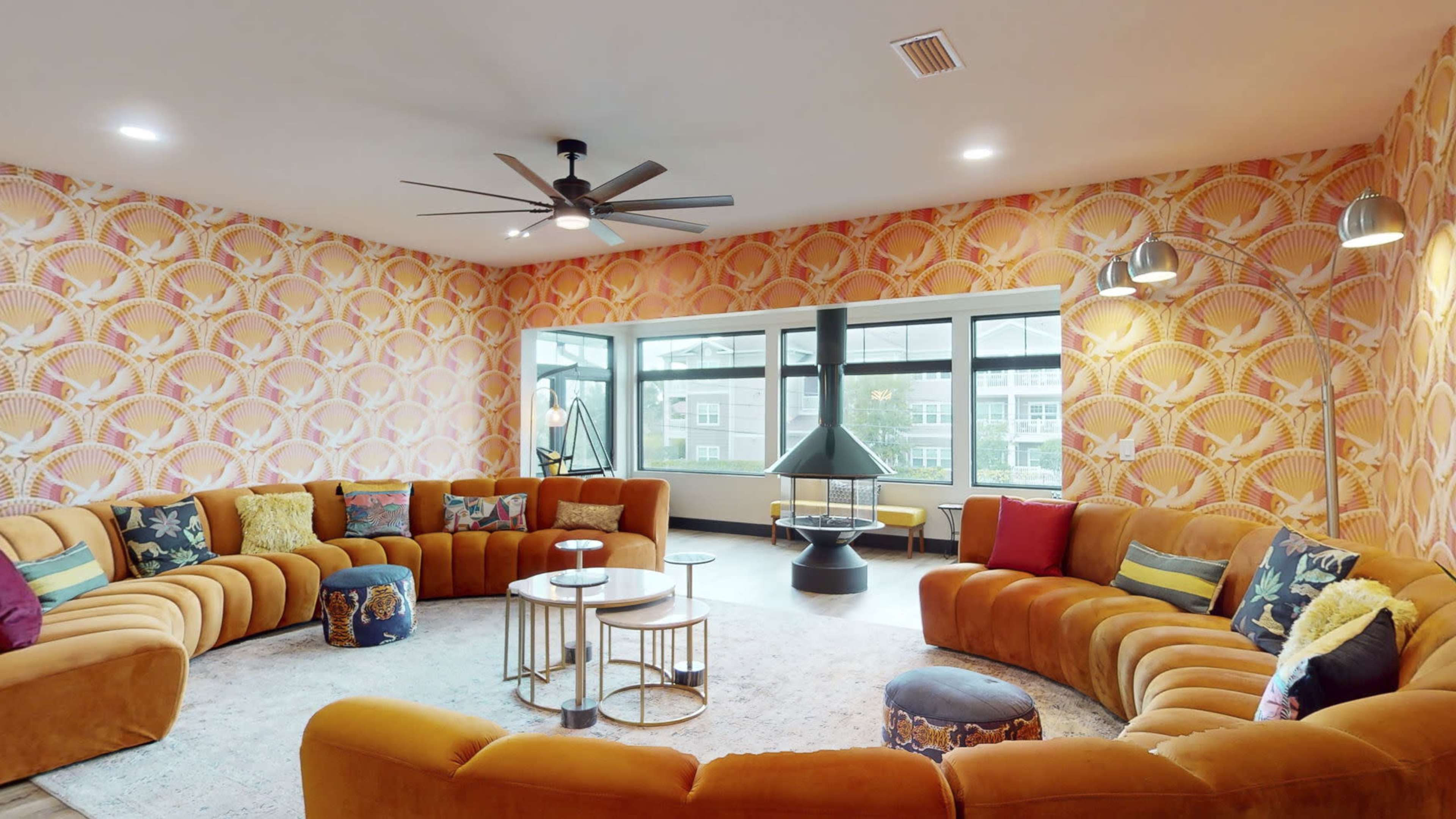 A spacious living room features a circular arrangement of orange upholstered sofas, colorful cushions, a coffee table set, and large windows allowing natural light.