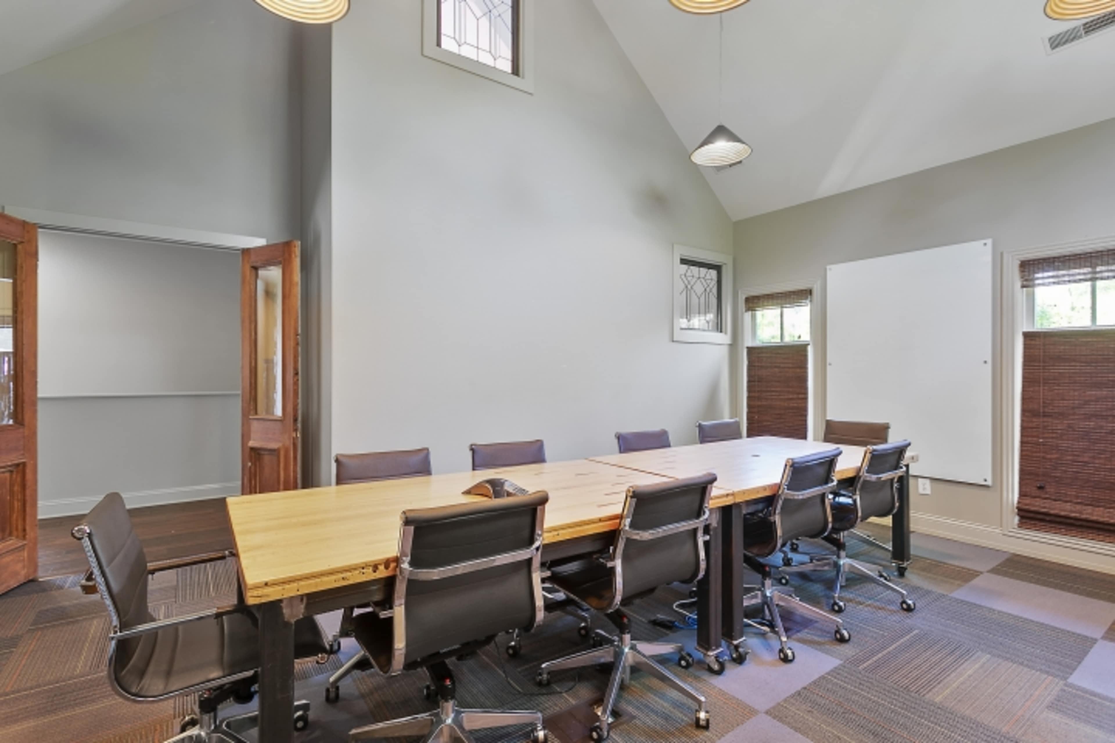 A conference room features a long wooden table surrounded by eight chairs, with large windows and a high ceiling.