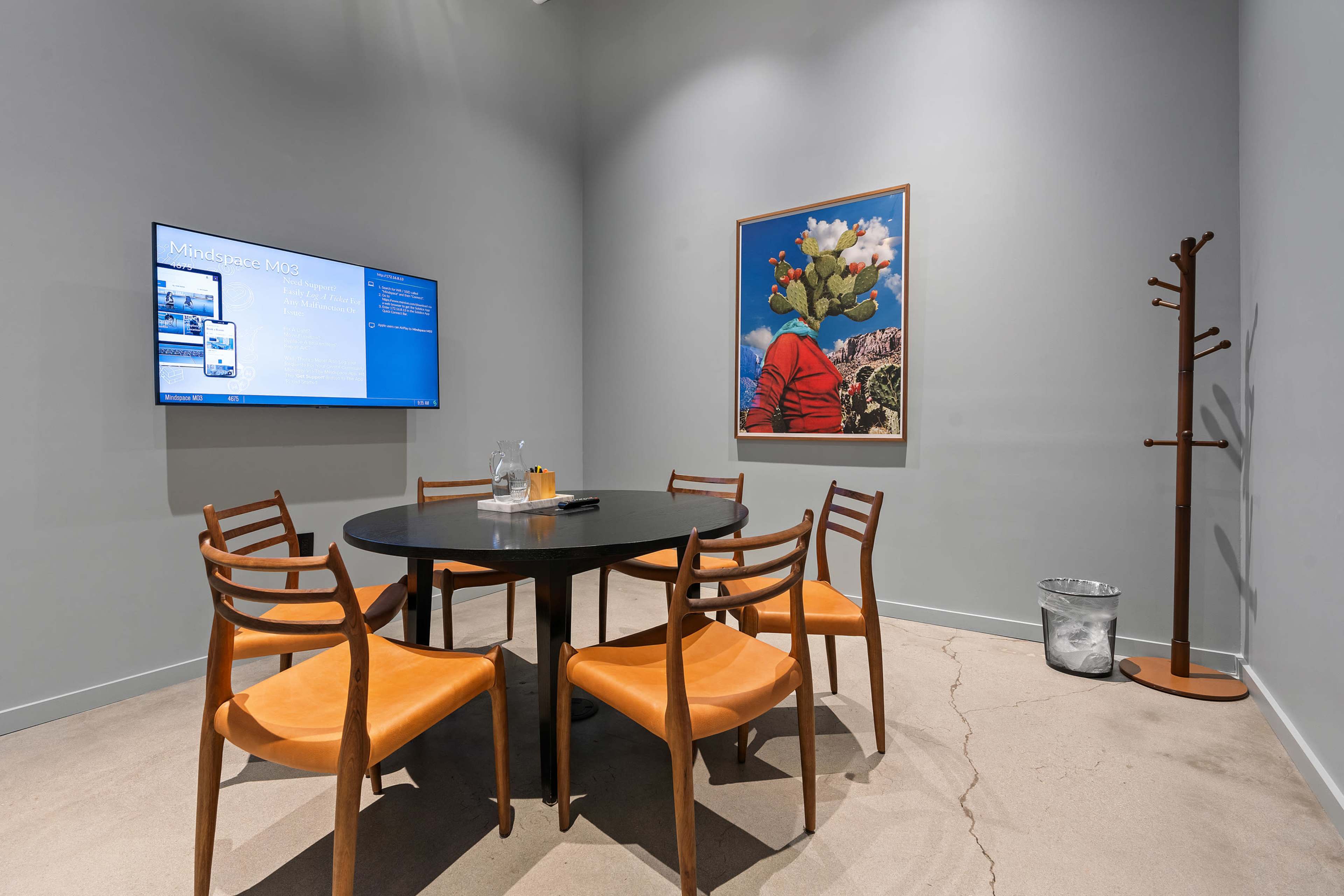 A modern meeting room features a round table surrounded by six wooden chairs, a wall-mounted screen displaying information, a framed artwork, and a coat rack in the corner.